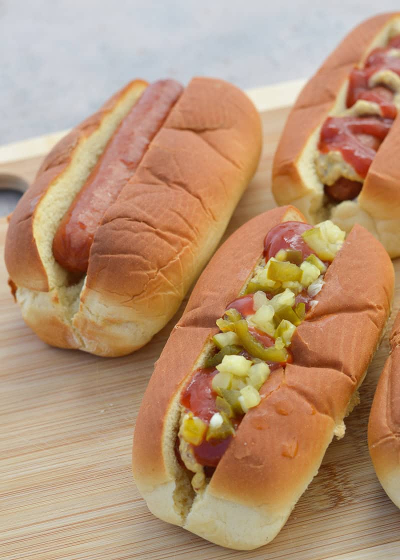 one plain hot dog, a hot dog with relish, and a hotdog with ketchup and mustard on a cutting board