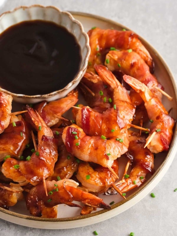 bacon wrapped shrimp, sprinkled with chives on a plate with a bowl of bbq sauce for dipping