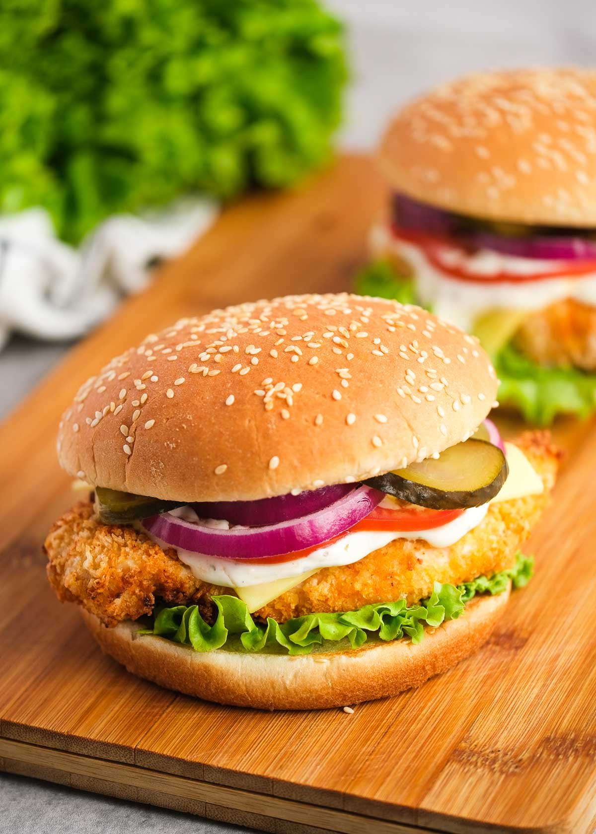 two loaded chicken sandwiches on a wooden cutting board with pickles, tomatoes, red onions, mayonnaise, cheese, and lettuce with a sesame bun