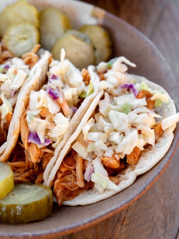 a plate loaded with sliced pickles and juicy bbq chicken tacos topped with coleslaw