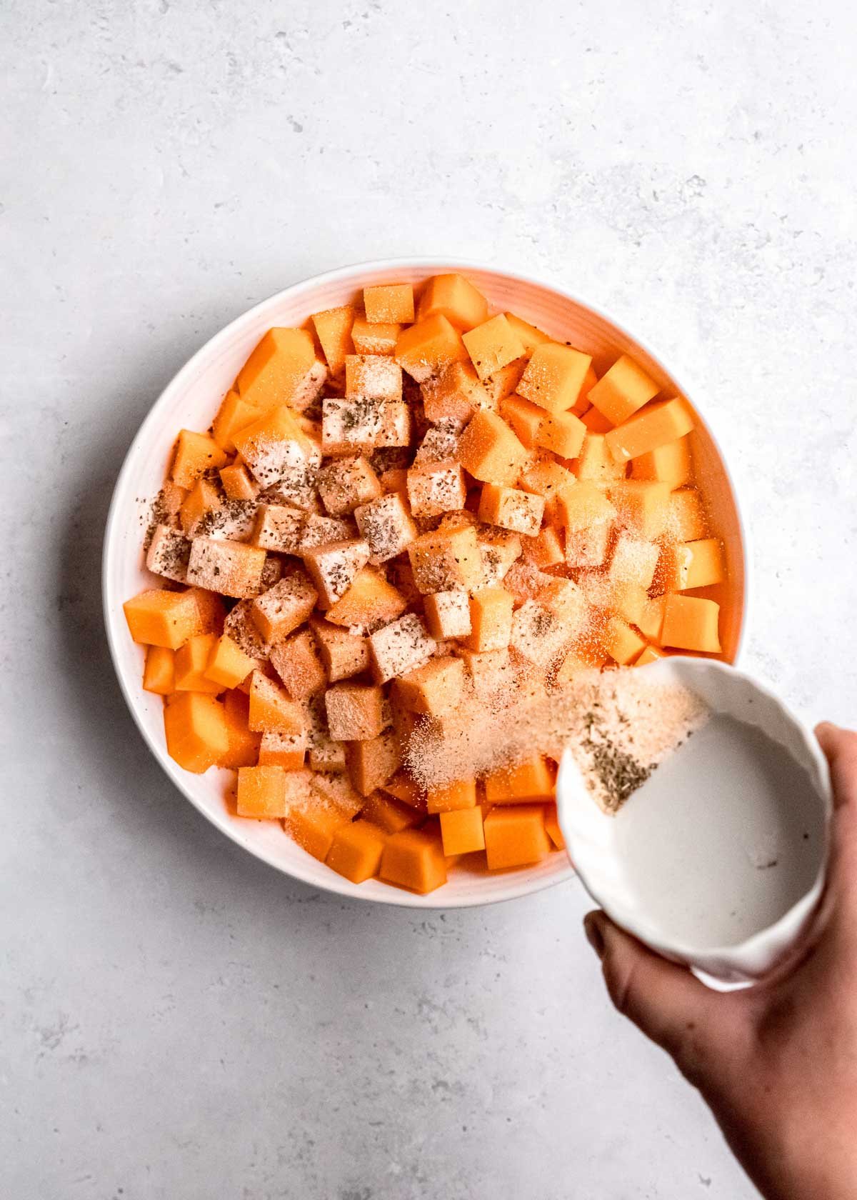 adding seasoning to cubed butternut squash in white bowl