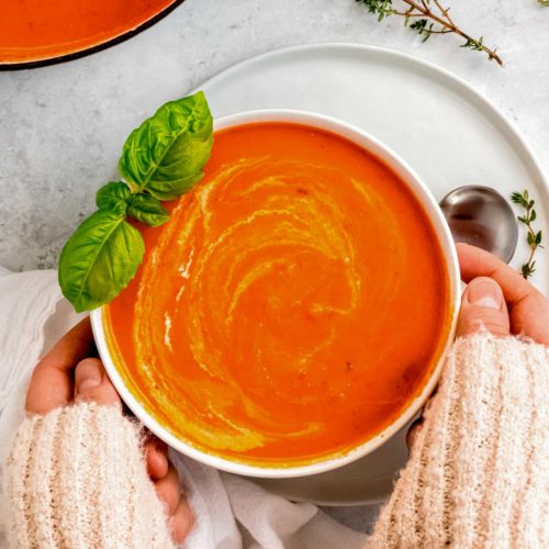 two hands grabbing a white bowl full of creamy roasted red pepper soup, garnished with basil, heavy cream, and thyme