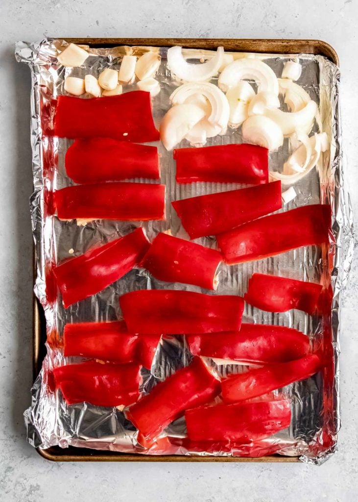 sliced red peppers, cut onion, and garlic cloves on a prepared baking sheet