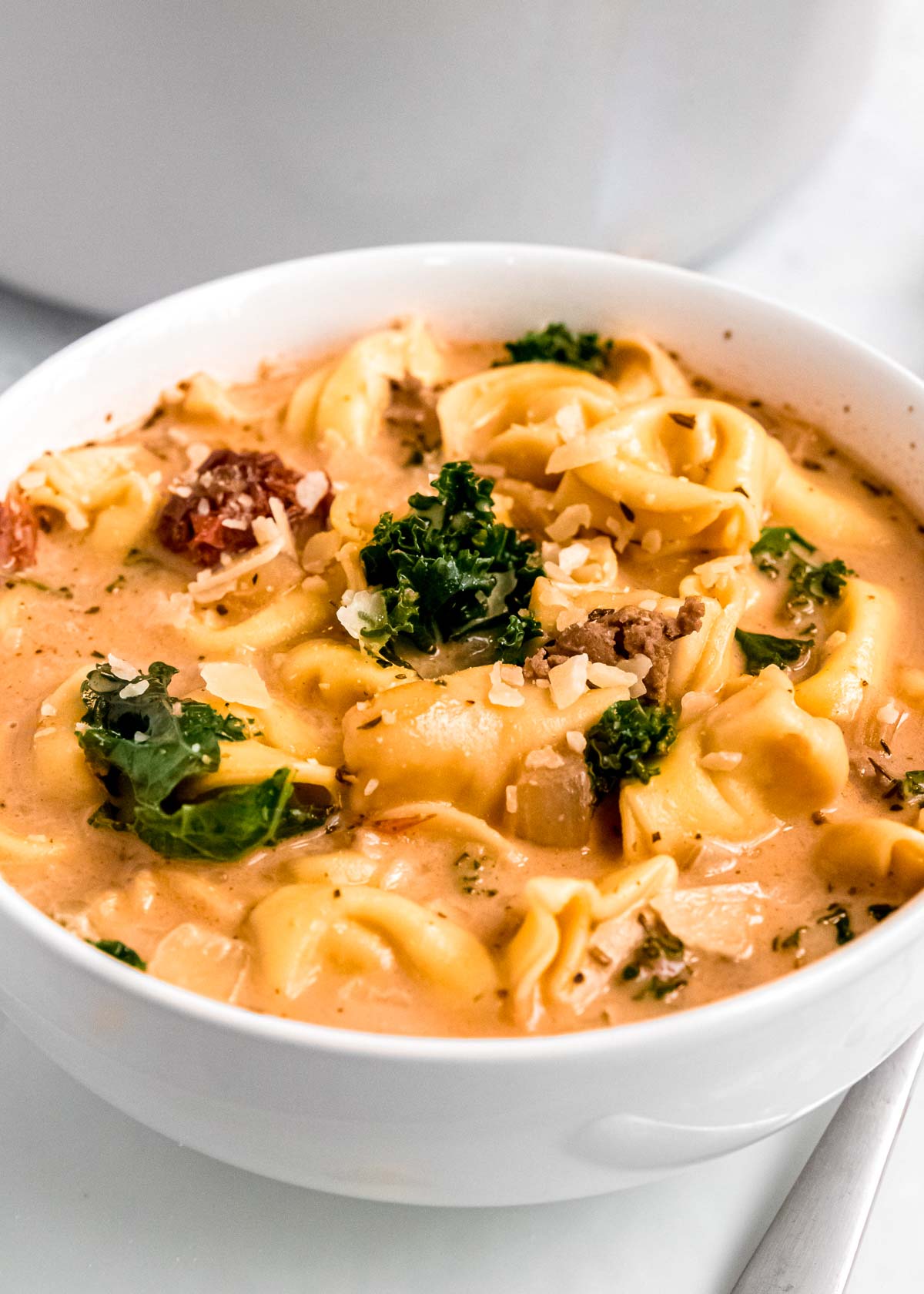 a white bowl full of creamy sausage tortellini soup with kale and sundried tomatoes