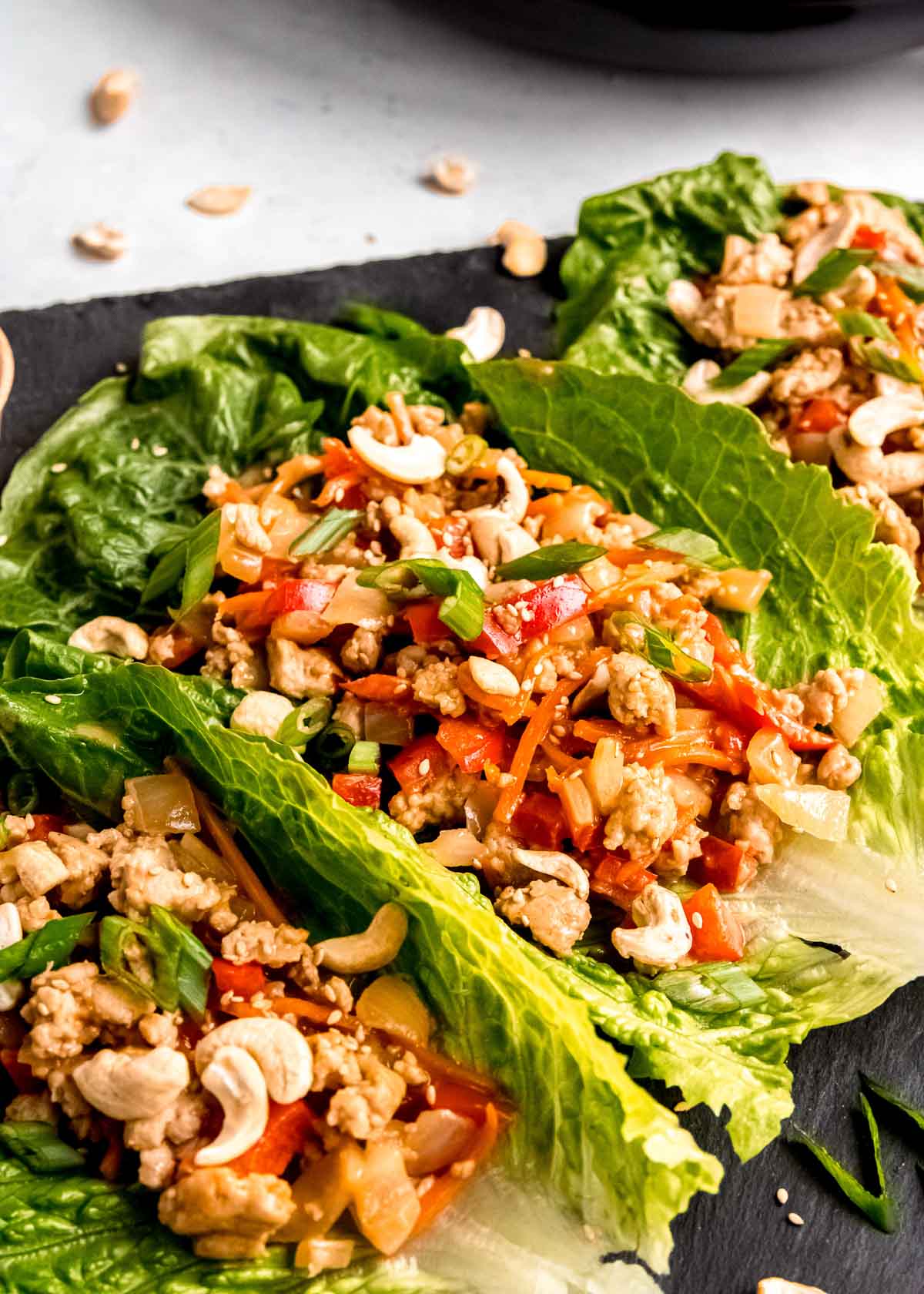 three lettuce wraps filled with thai chicken, peppers, and carrots and topped with cashews, green onions, and sesame seeds.