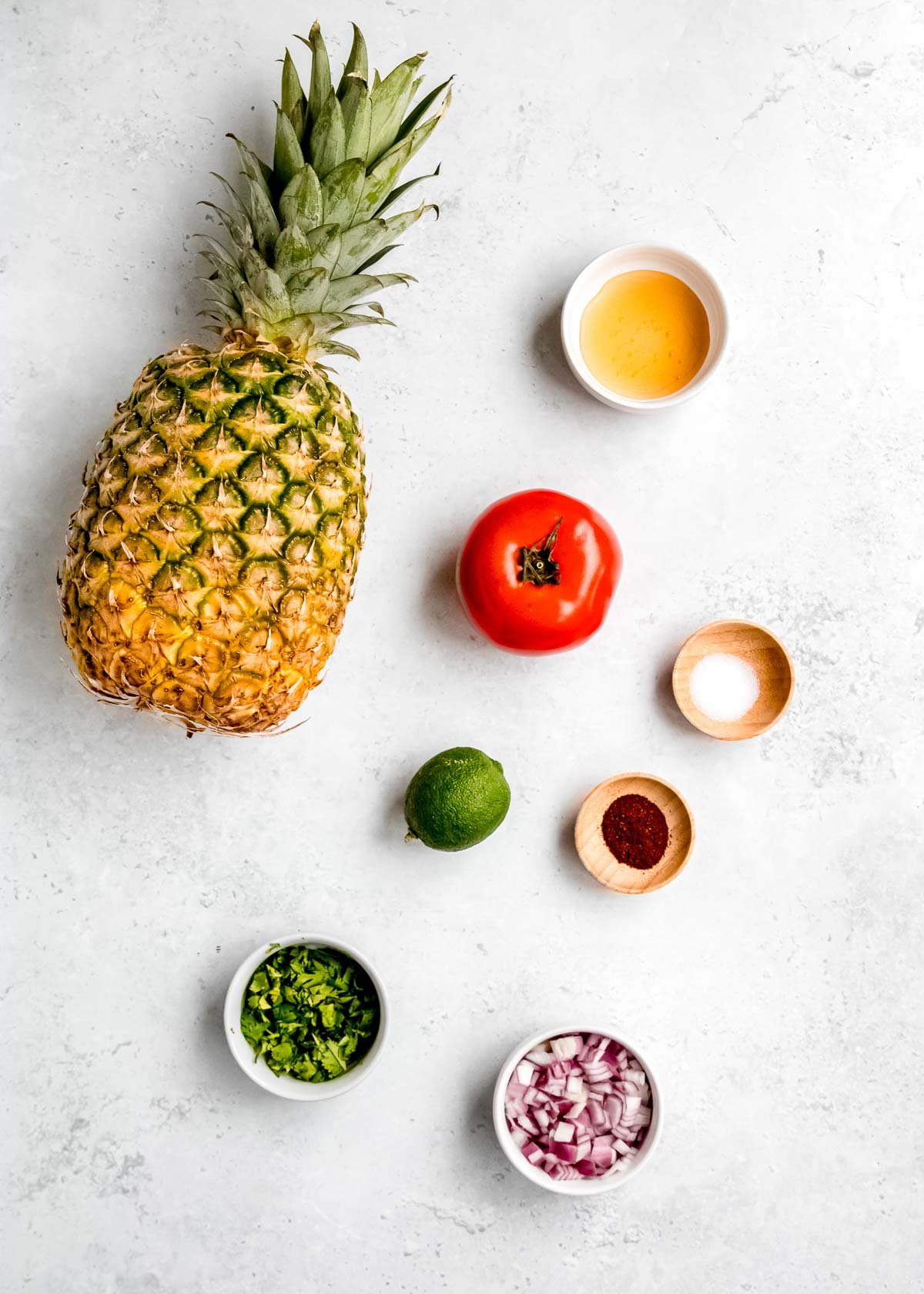 ingredients for pineapple salsa, including a fresh pineapple, tomato, lime, apple cider vinegar, salt, chili powder, chopped red onion, and chopped cilantro