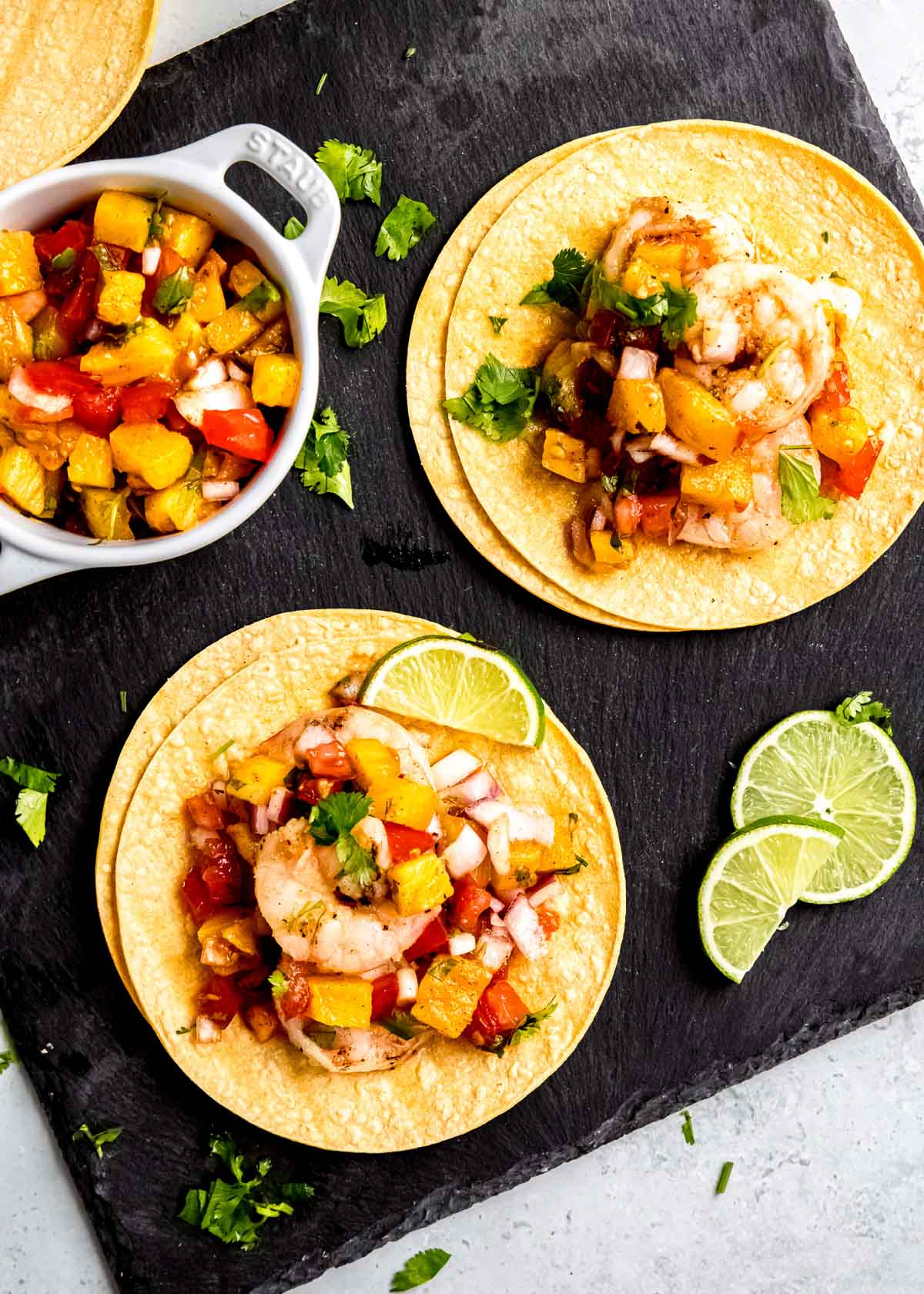 faraway shot of serving board topped with a bowl of grilled pineapple salsa, chopped cilantro, lime wedges, and two grilled shrimp tacos