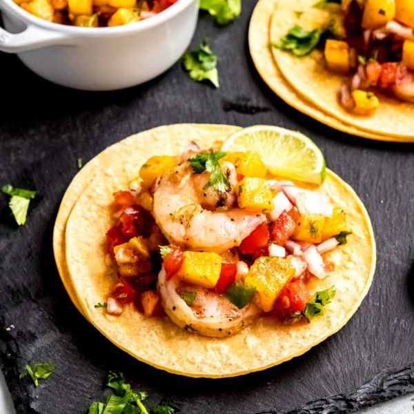 two grilled shrimp tacos on a black serving board, surrounded with pineapple salsa, cilantro, and lime wedges