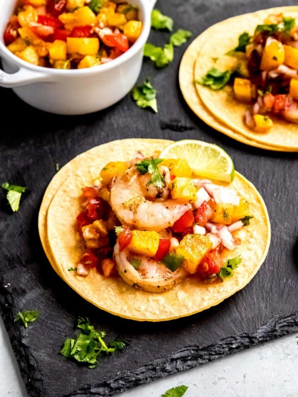 two grilled shrimp tacos on a black serving board, surrounded with pineapple salsa, cilantro, and lime wedges