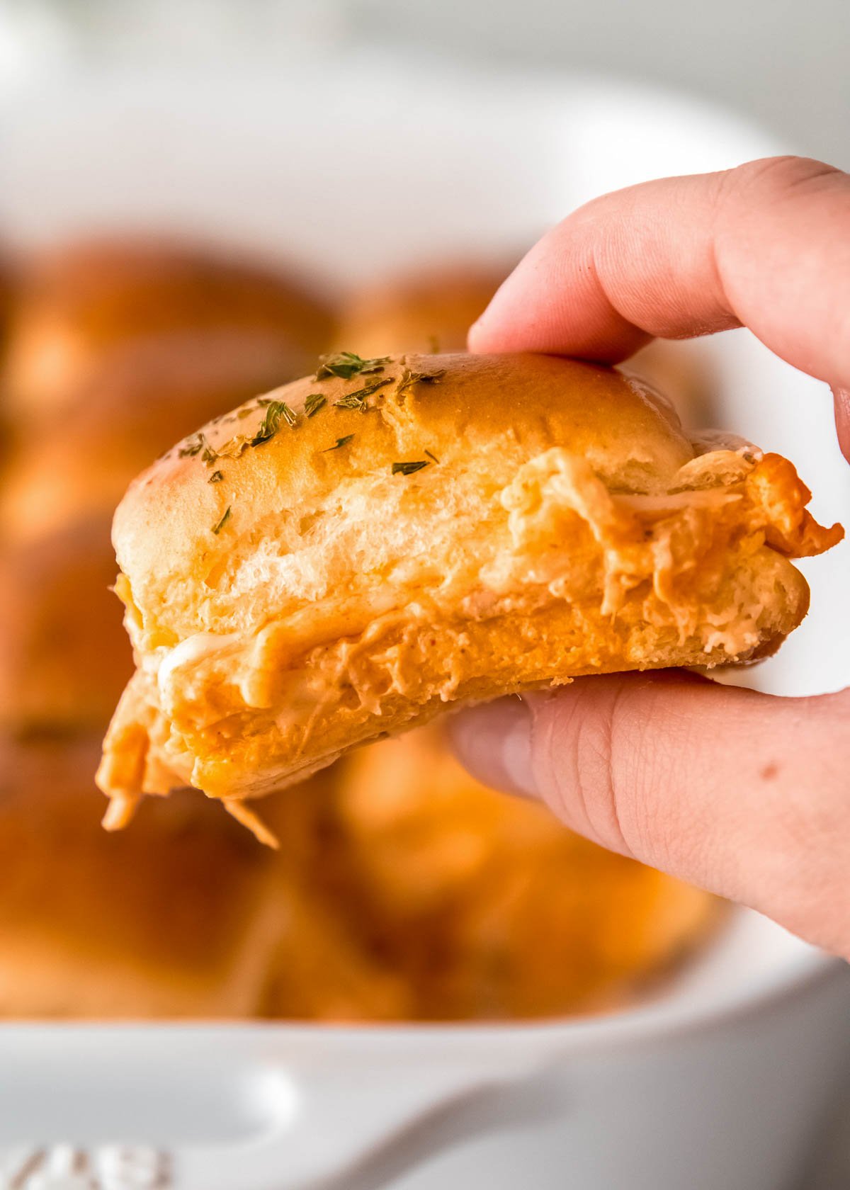 a hand holding a cheesy buffalo chicken slider in front of a pan full of easy party sliders