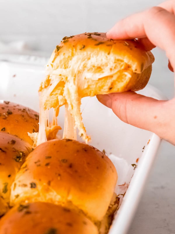 a hand grabbing a buffalo chicken slider from a casserole dish; gooey, melted cheese strings down