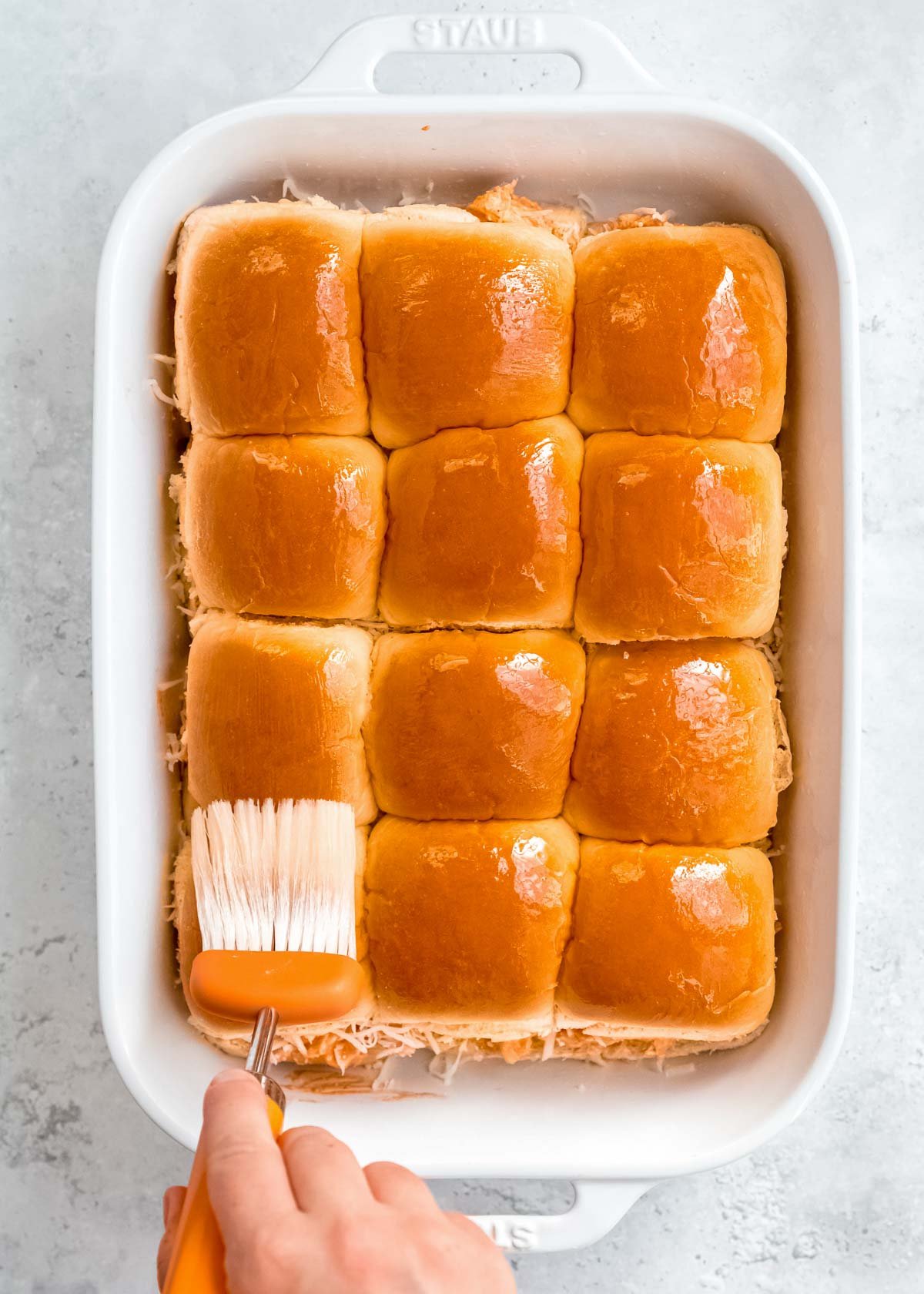 top buns added to the white casserole dish; a hand using a brush to coat the top of the buns with melted butter