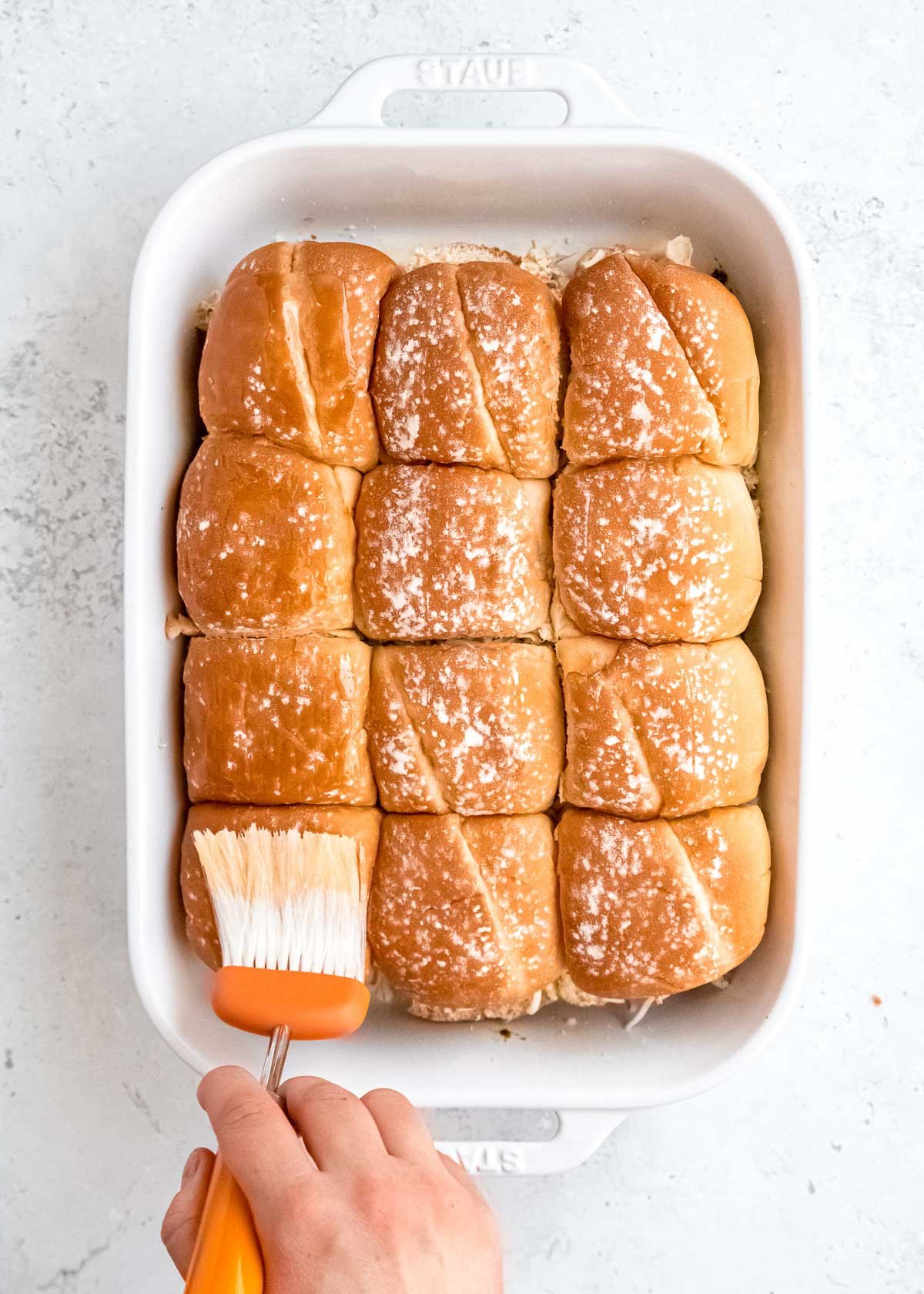 the tops of the buns added to the sliders; a hand using a culinary brush to spread butter over the buns