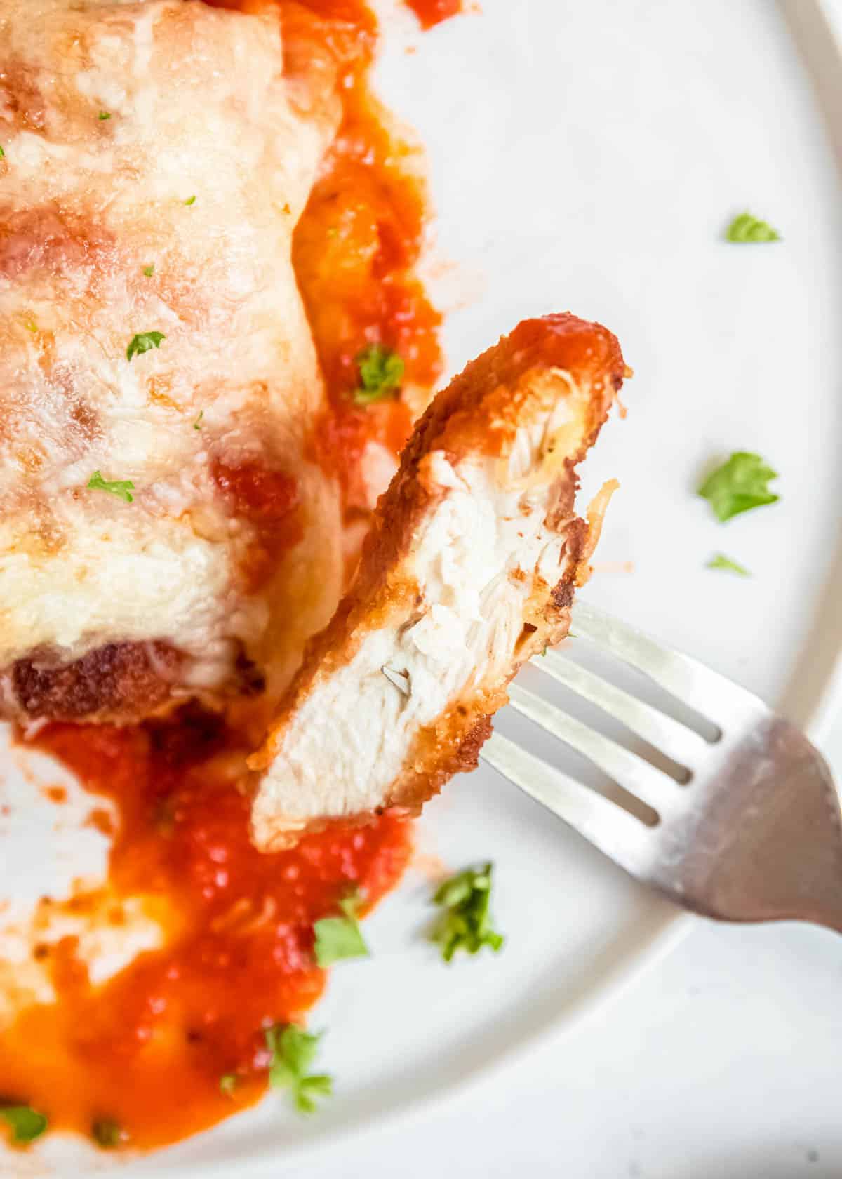 a single bite of chicken parmesan on fork with chicken breast and plate in background