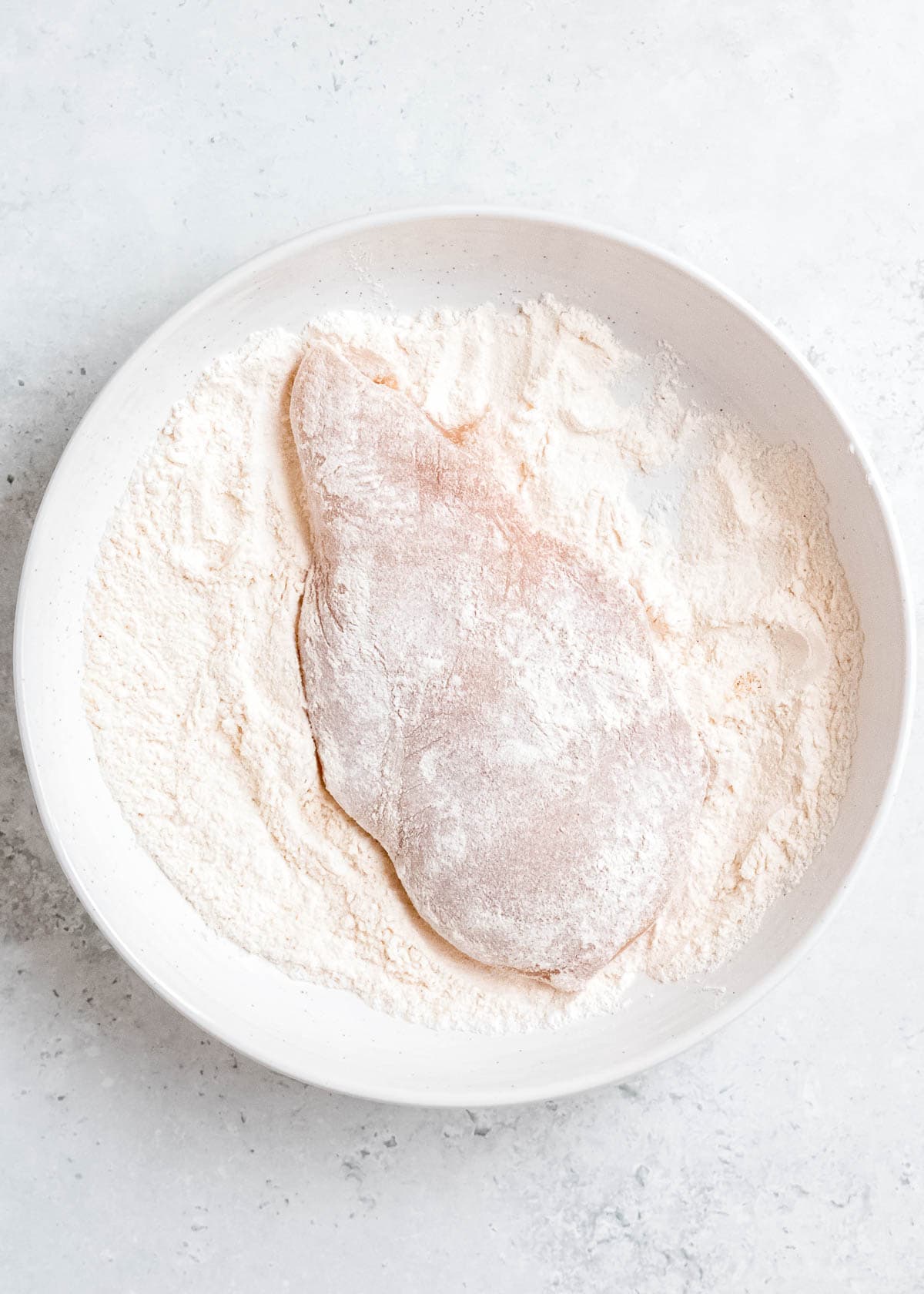 chicken breast being rolled in flour in white bowl