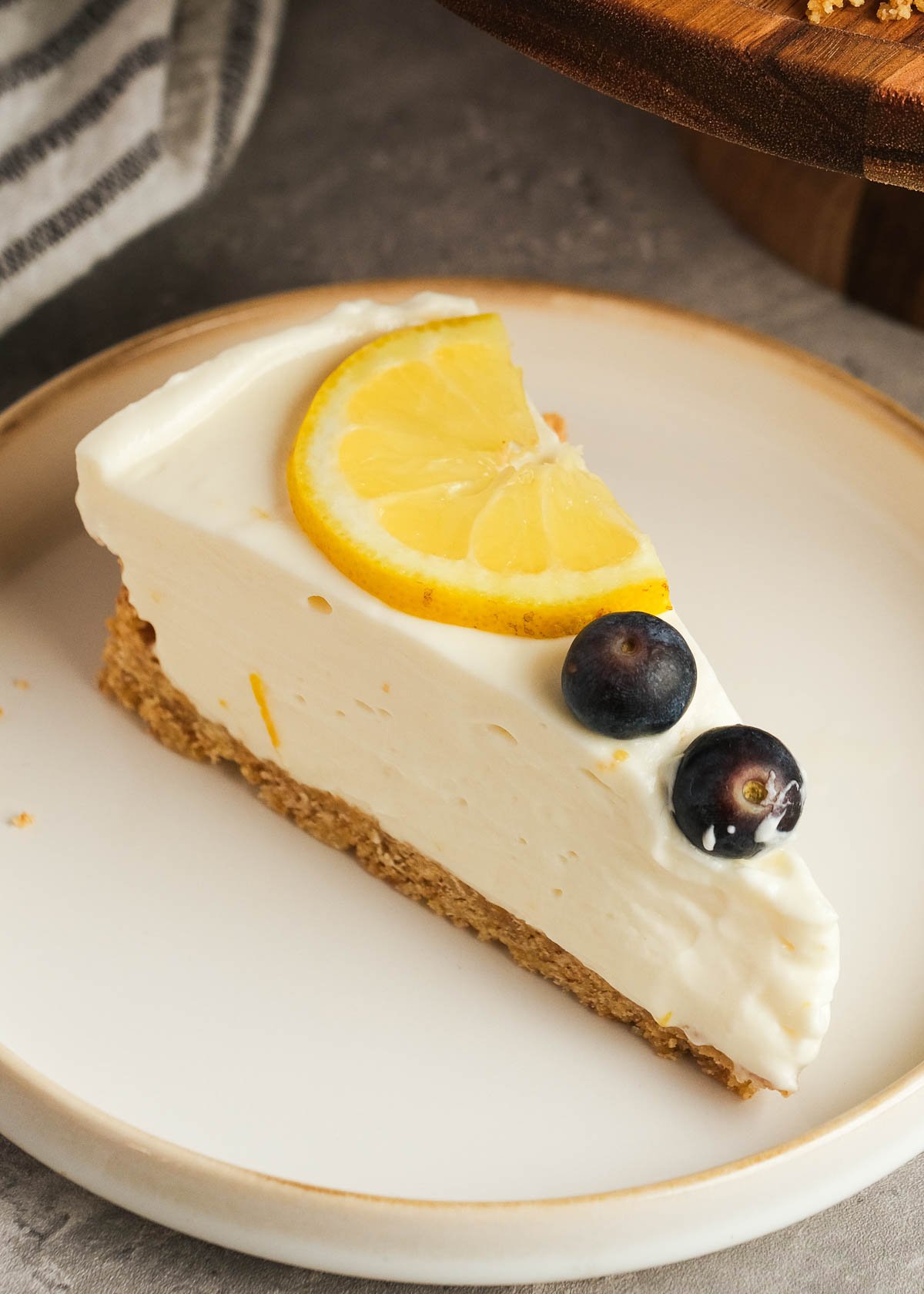 a white plate topped with a perfect slice of easy no-bake lemon cheesecake garnished with lemon slices and fresh blueberries