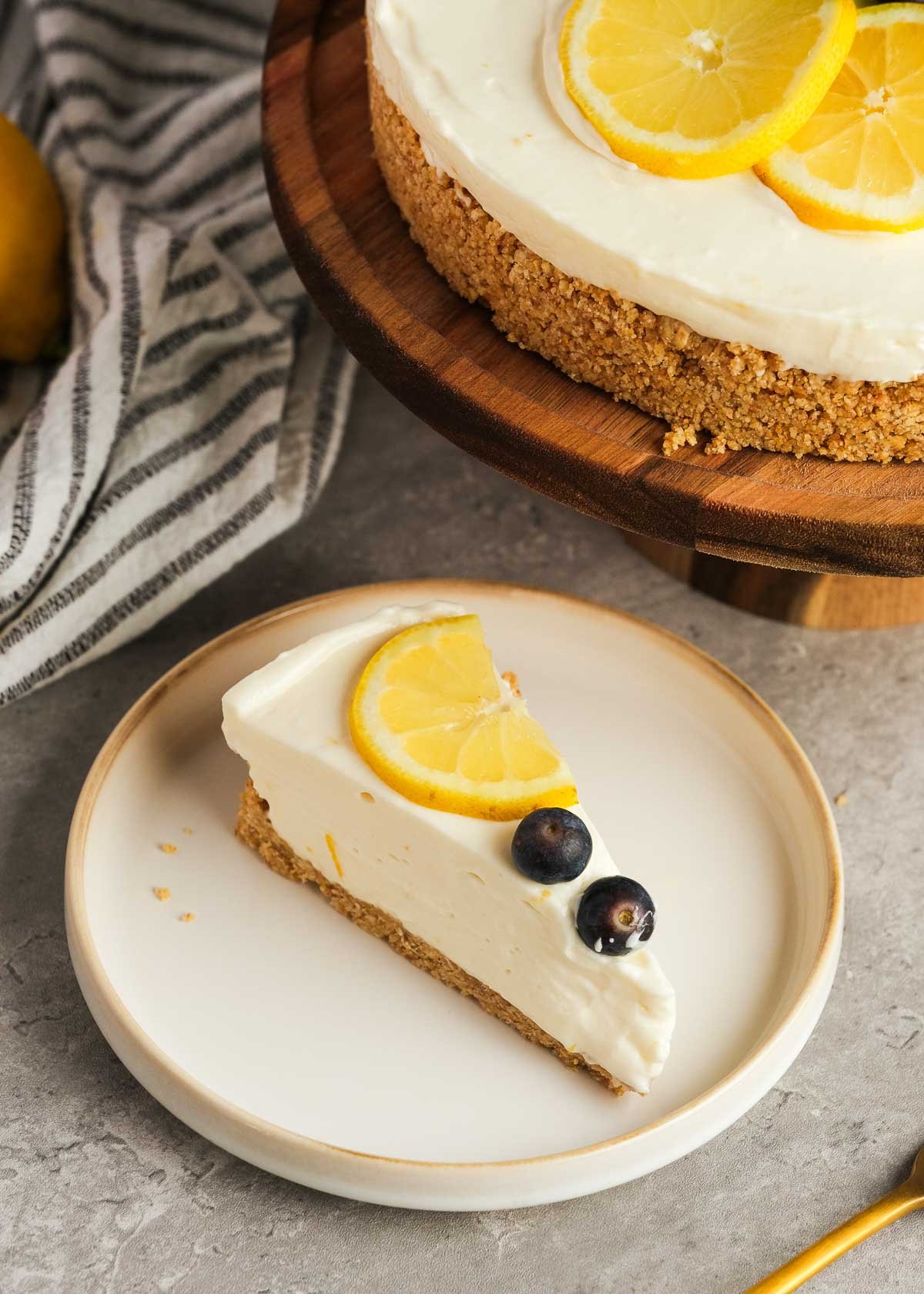 a white plate topped with a slice of no bake lemon cheesecake topped with a lemon slice and two blueberries; in the background there is the remaining cheesecake and a dish towel