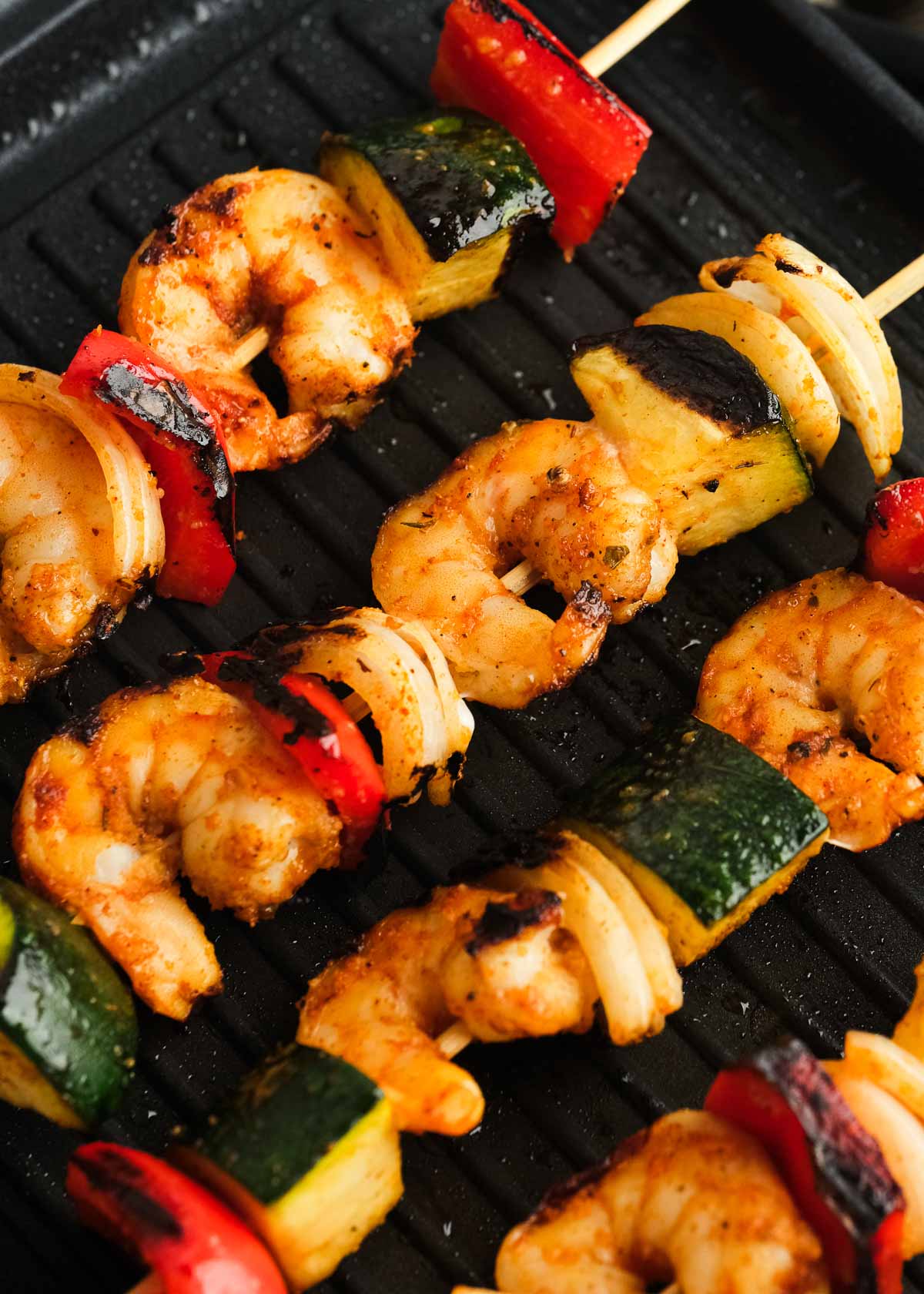 closeup view of four skewers filled with shrimp, zucchini, bell peppers, and onion on the grill