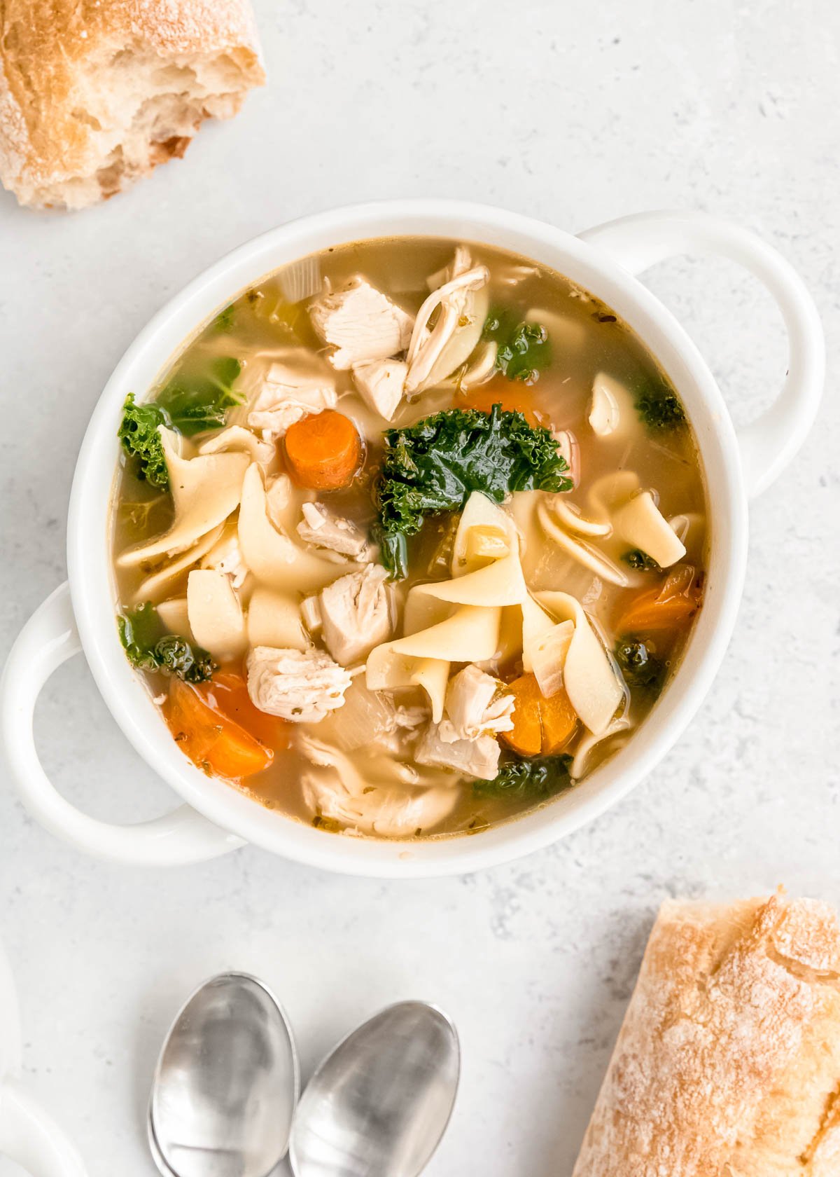 overhead shot of a white bowl filled with classic chicken noodle soup with egg noodles. bread and spoons shown in the background