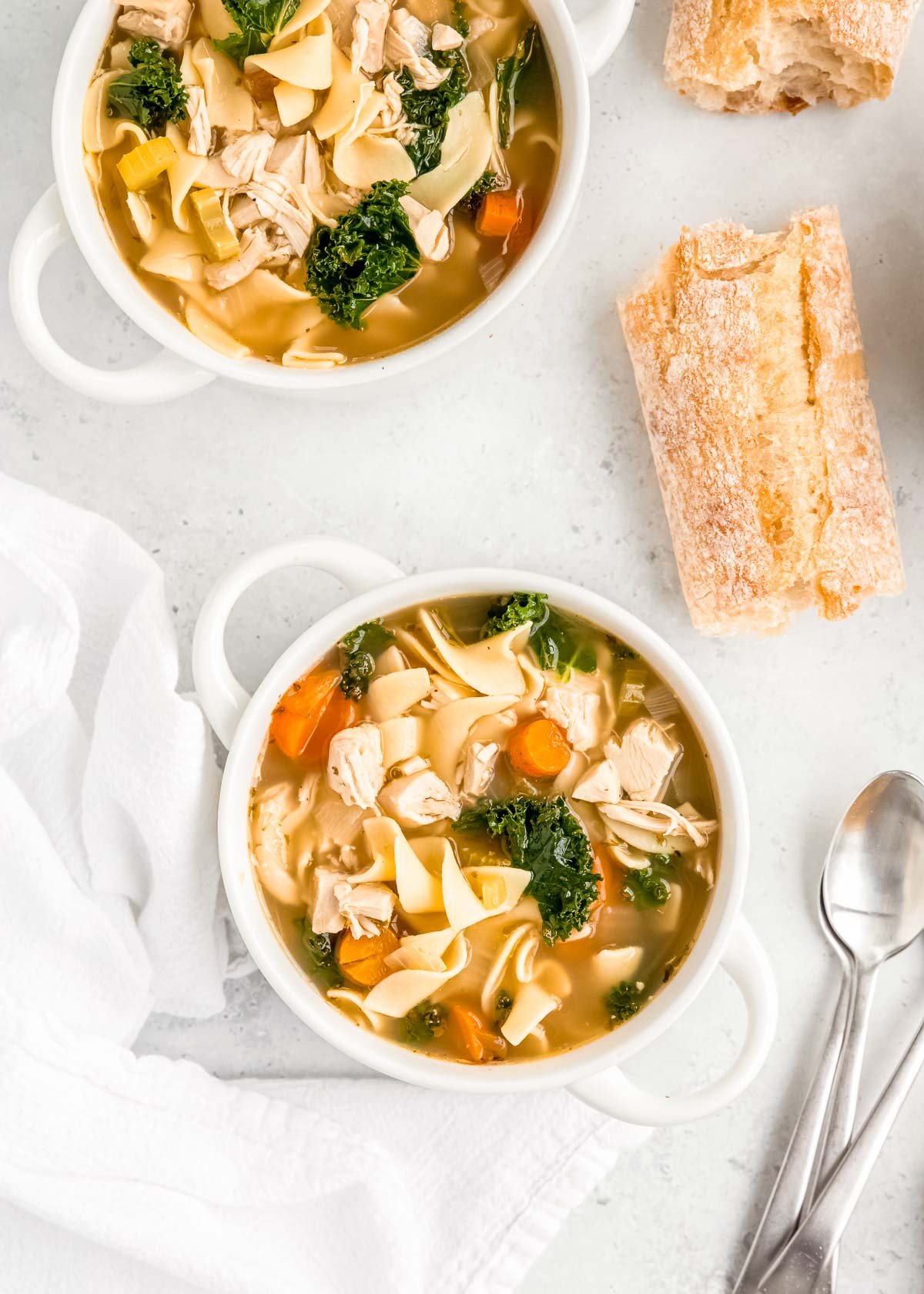 two bowls of chicken noodle soup with egg noodles and kale with white napkins, silver spoons, and two pieces of bread on a white table