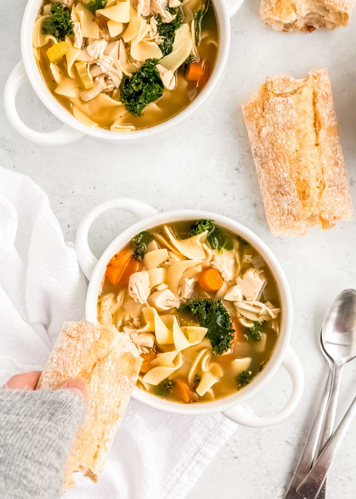 crusty bread dipping into a bowl of simple chicken noodle soup with egg noodles