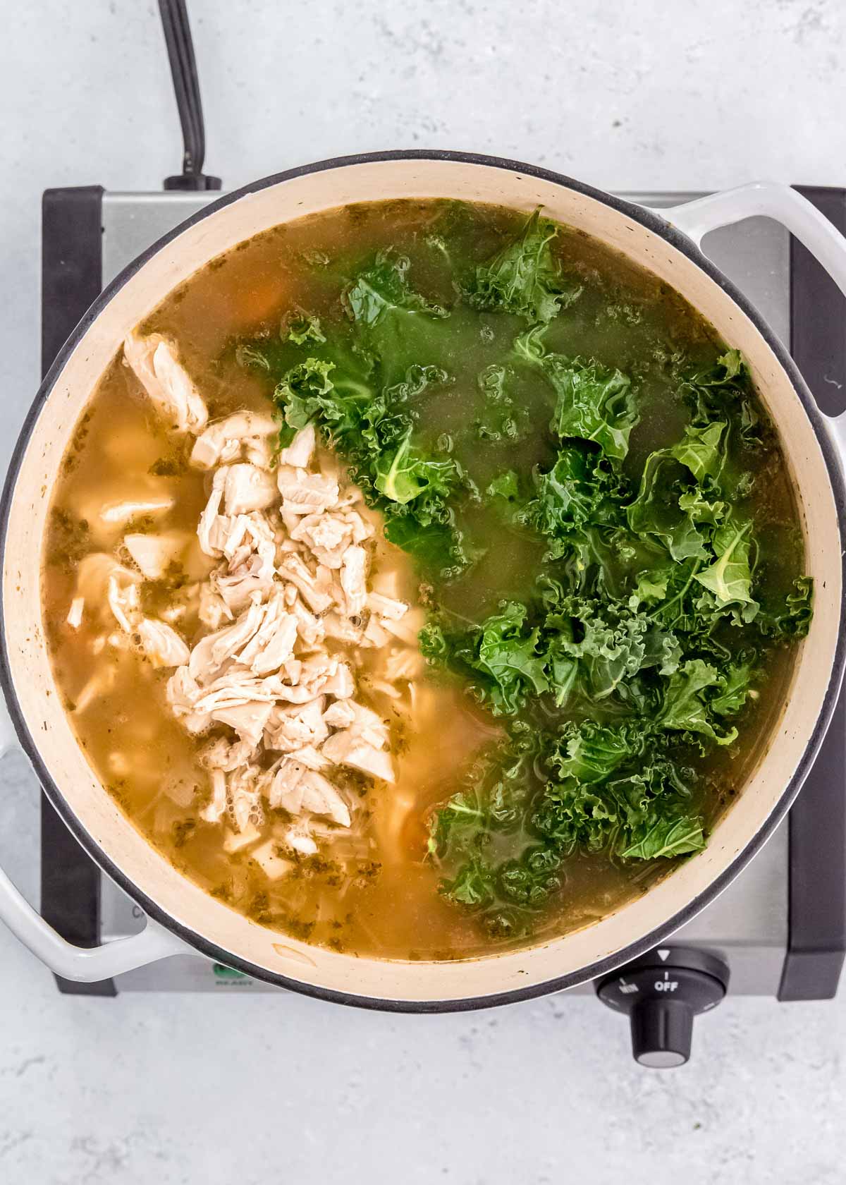 shredded chicken and chopped kale added into a large pot filled with broth, egg noodles, and vegetables