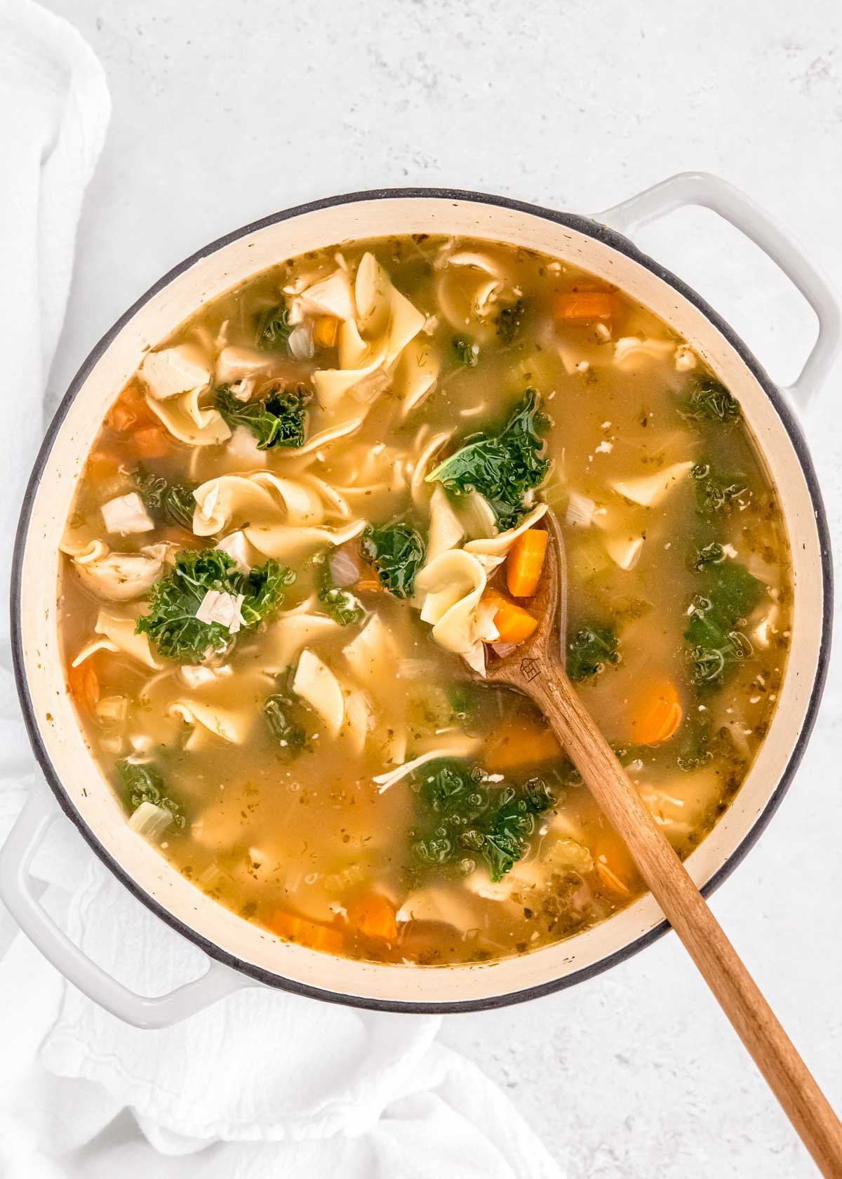 overhead shot of a large pot of chicken noodle soup with a wooden spoon picking up egg noodles, shredded chicken, carrots, and kale in a delicious broth.