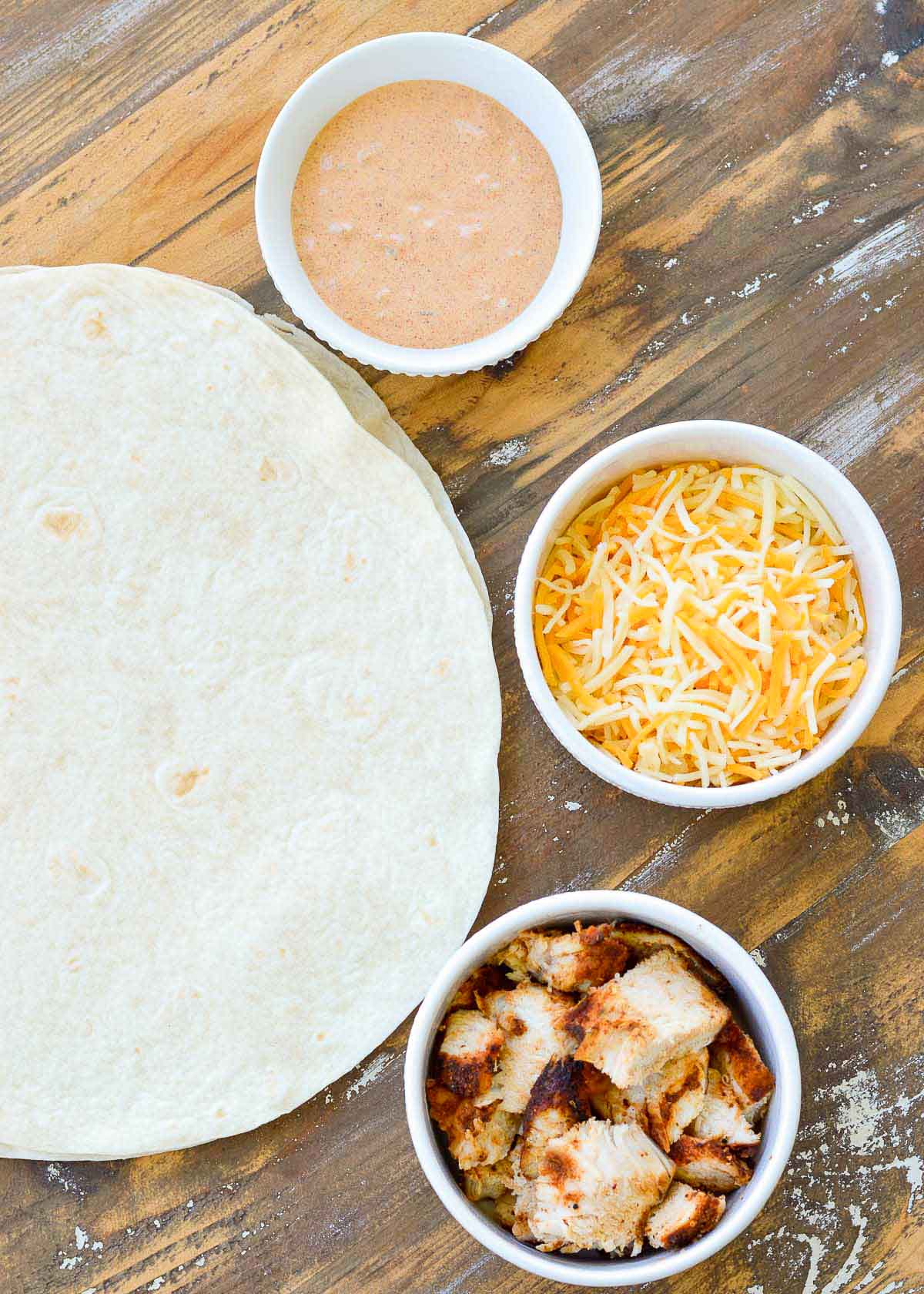 tortillas on a wooden counter with ramekins of sliced chicken, shredded cheese, and creamy jalapeno sauce