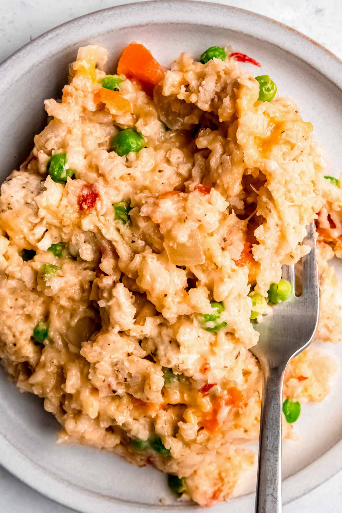 overhead shot of cheesy chicken and rice with peas and carrots on a white plate; fork on side