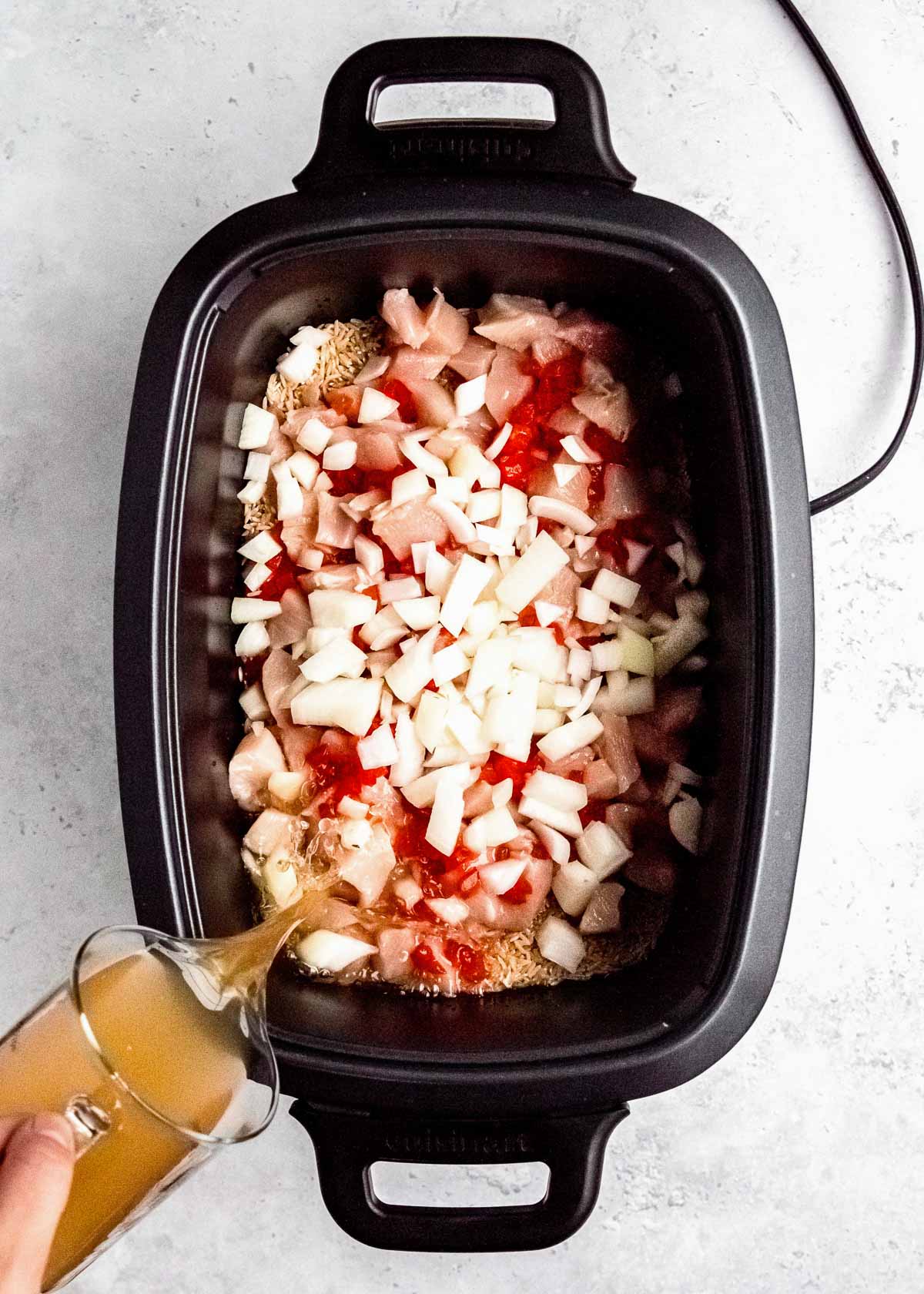 chicken broth being poured into black crockpot filled with cubed chicken, chopped onions, pimentos, and brown rice