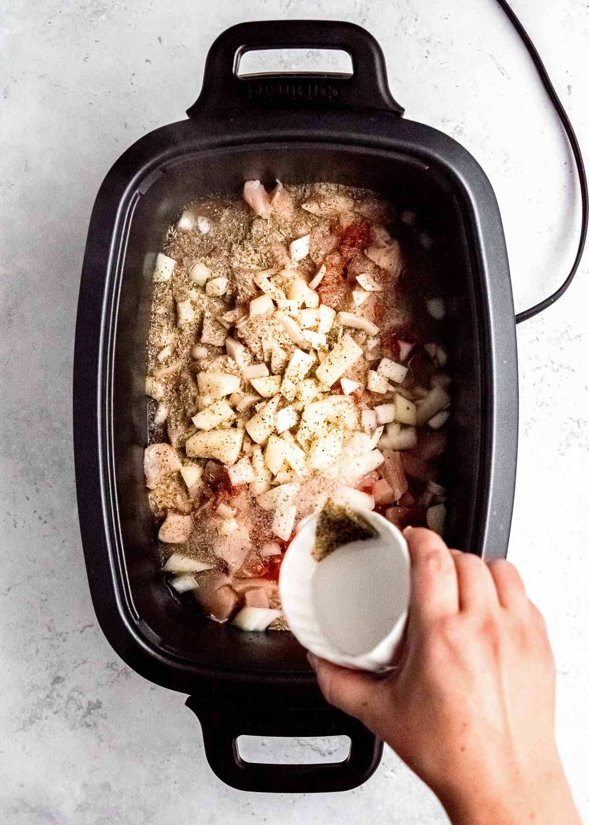 seasonings being sprinkled over cubed chicken, brown rice, chopped onions, pimentos, and chicken stock in a crockpot