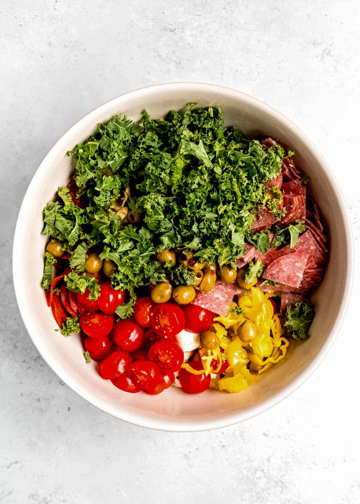 kale, tomatoes, pepperoncini, olives, and cured meats added to a bowl with pasta