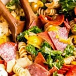 a giant bowl filled with antipasto pasta salad tossed with italian dressing with two wooden spoons