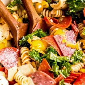 a giant bowl filled with antipasto pasta salad tossed with italian dressing with two wooden spoons