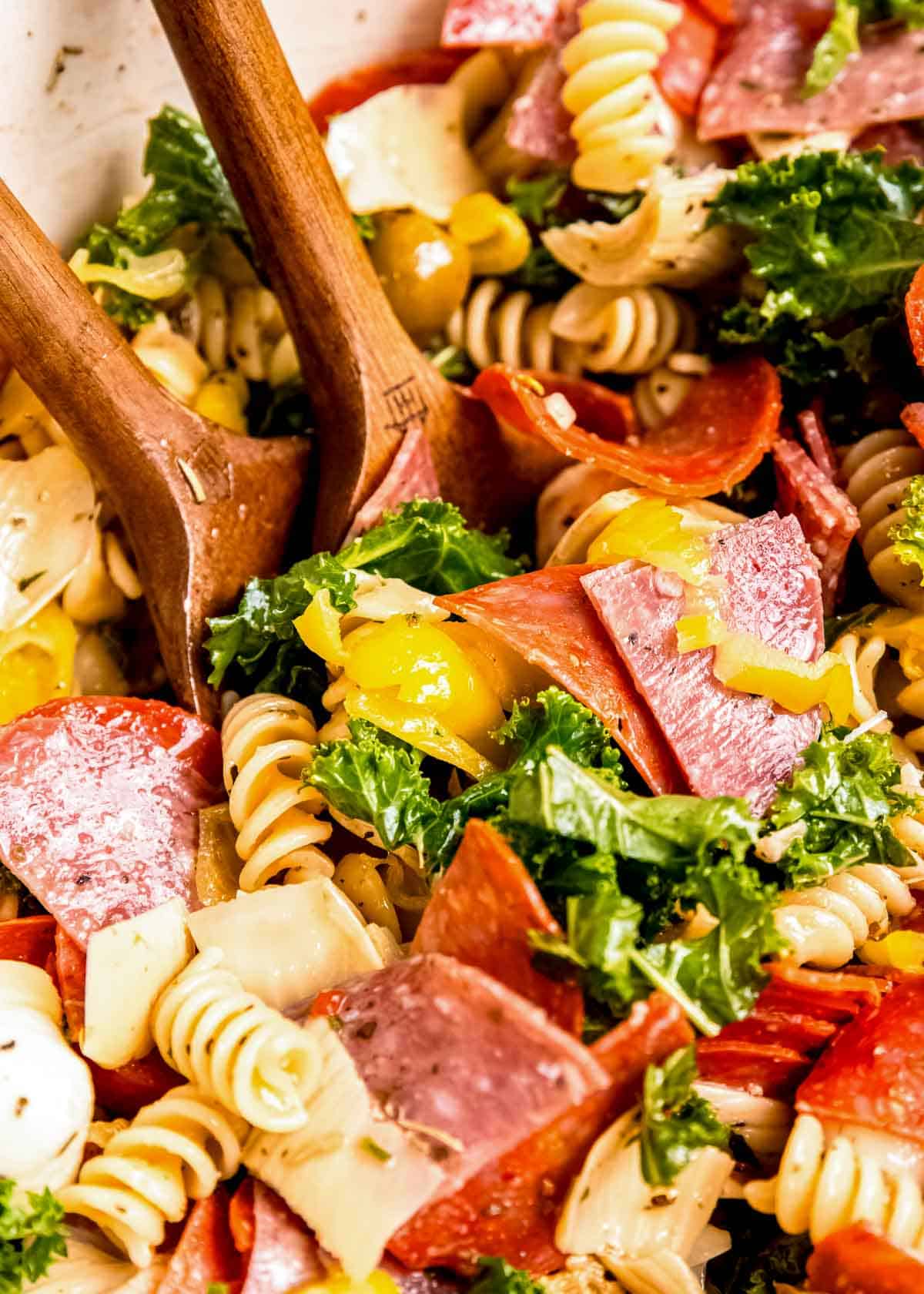 a giant bowl filled with antipasto pasta salad tossed with italian dressing with two wooden spoons