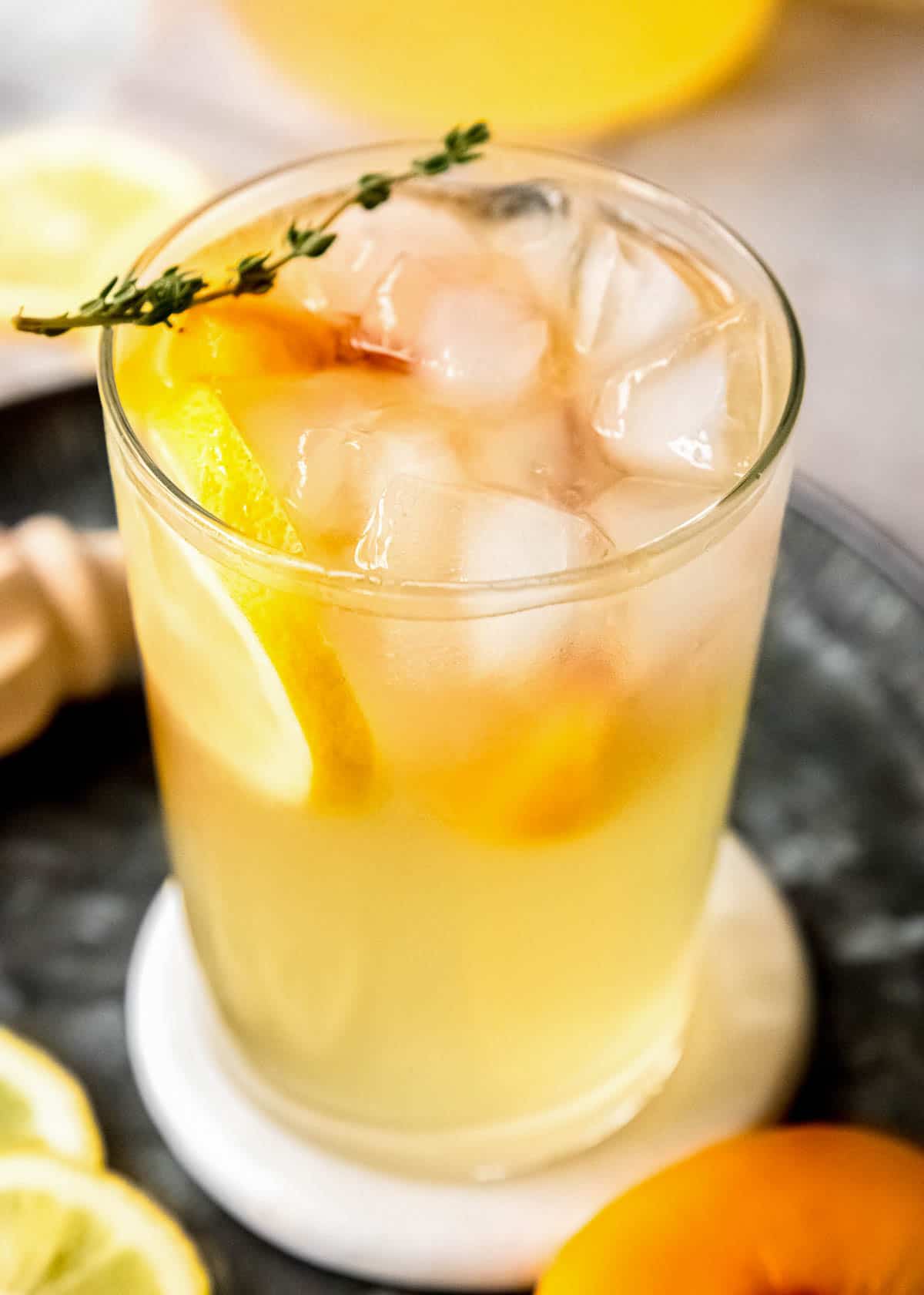 A glass of peach bourbon lemonade on ice, garnished with lemon and peach slices and a spring of thyme