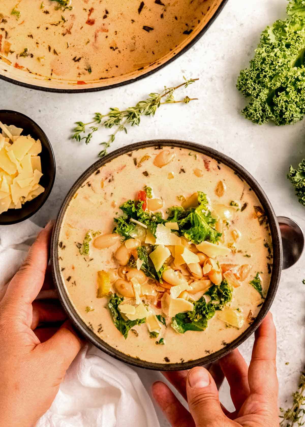 two hands picking up a big bowl of creamy tuscan soup with white beans, kale, parmesan cheese, and fresh herbs