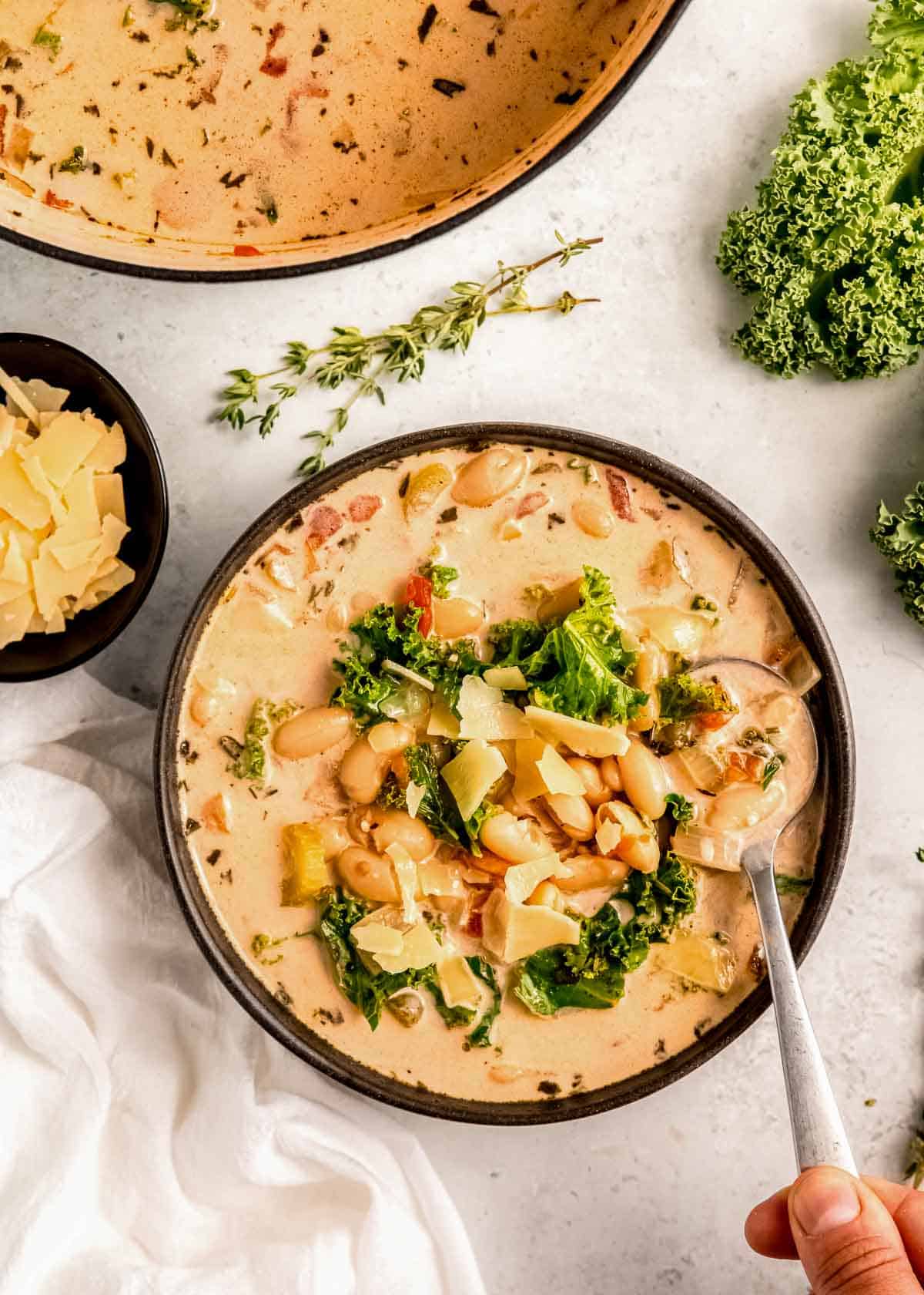 A bowl of creamy Tuscan white bean soup garnished with parmesan; spoon full of a nutritious bite