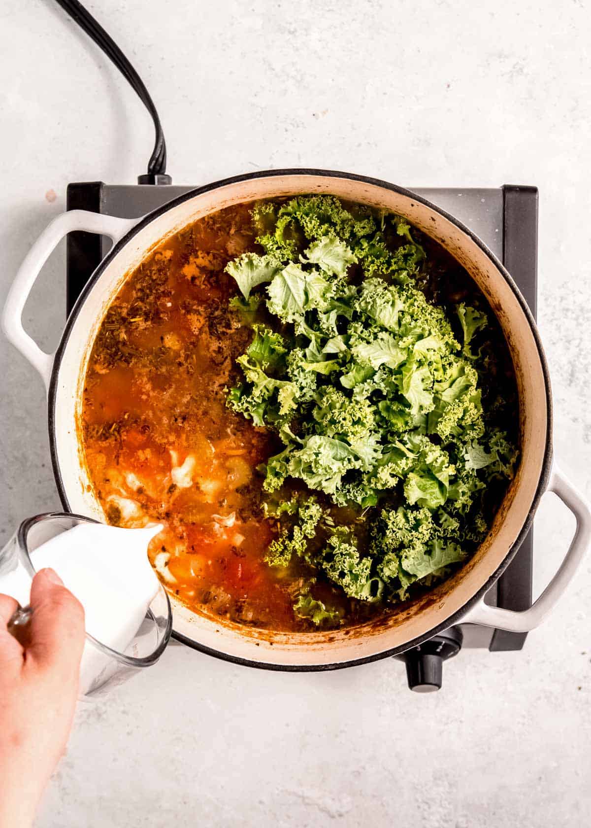 chopped kale and heavy cream being added to white bean soup in a dutch oven