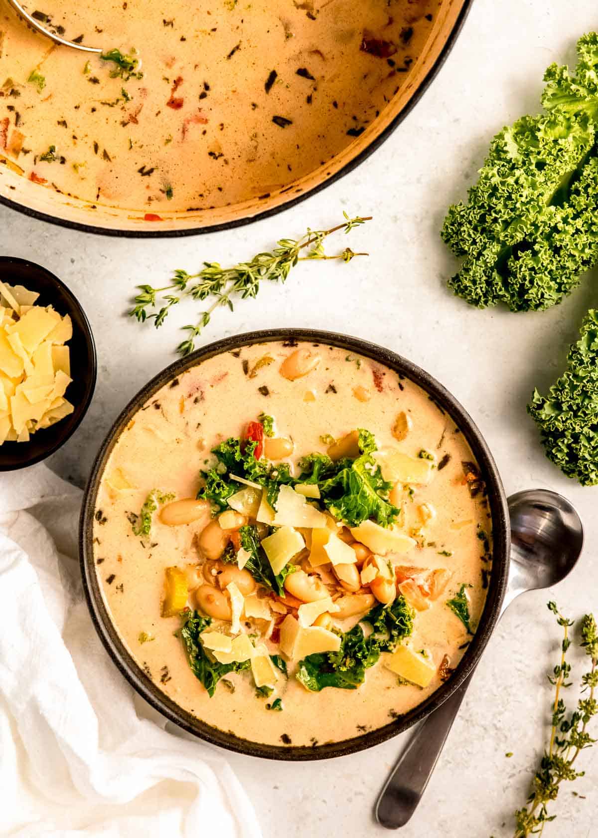 white countertop with a black bowl of tuscan white bean soup with fresh thyme, kale, and a side of parmesan