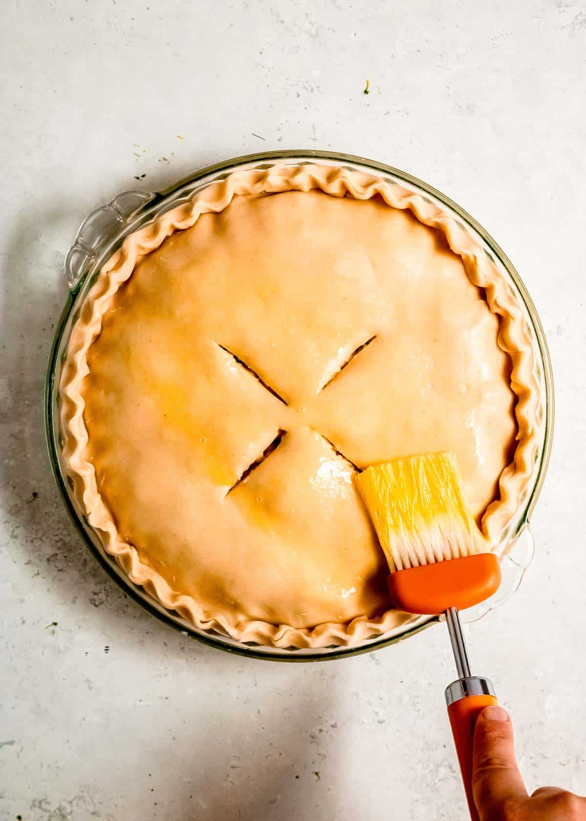 Egg wash being brushed onto the top crust of a chicken pot pie after the edges have been sealed and the venting slits have been cut