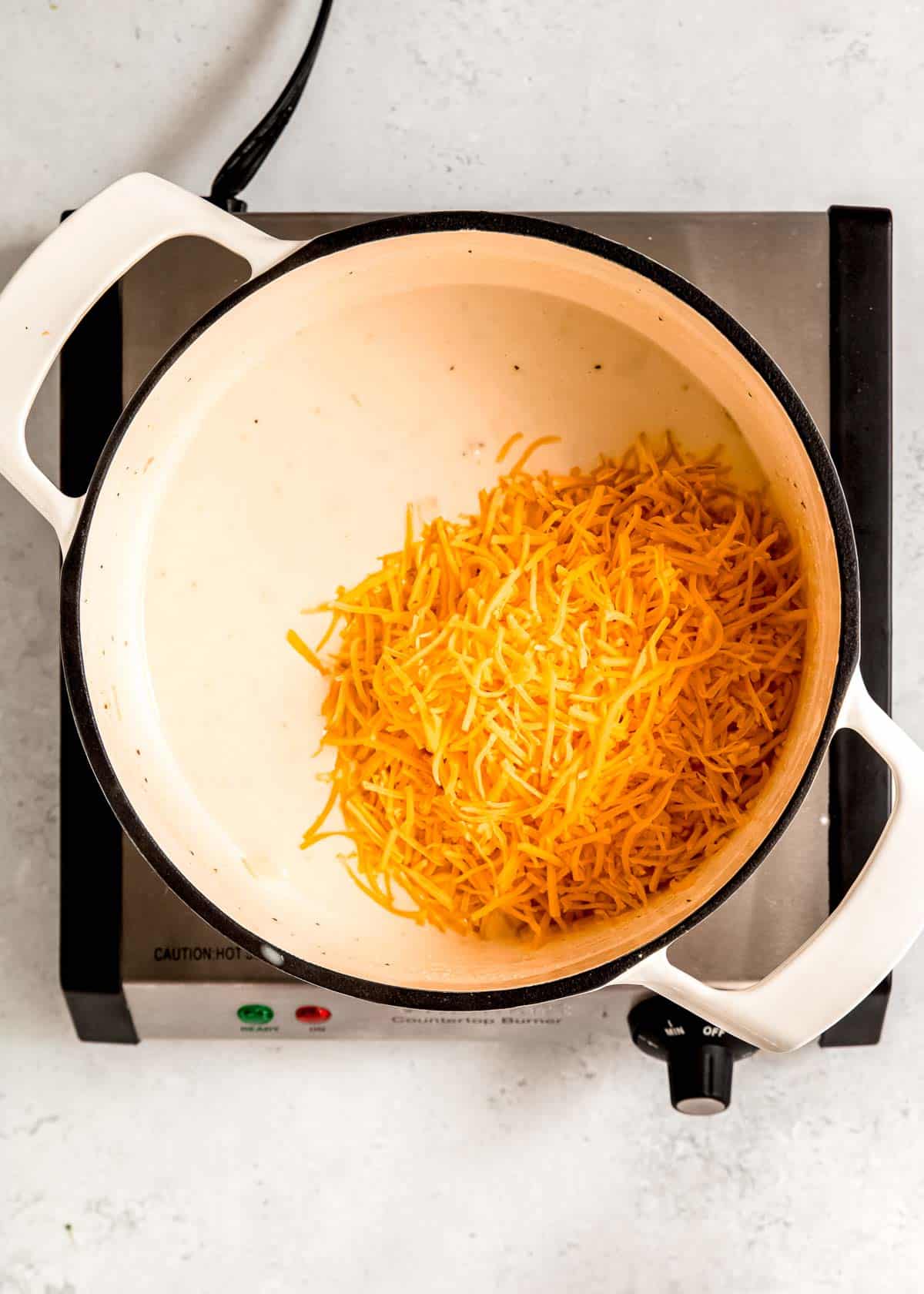 shredded cheddar cheese being added to the cream sauce in a large pot