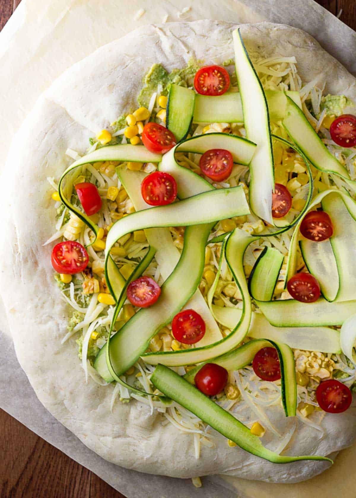 a parbaked flatbread pizza topped with cherry tomatoes, zucchini ribbons, charred corn, shredded mozzarella, and an avocado mash