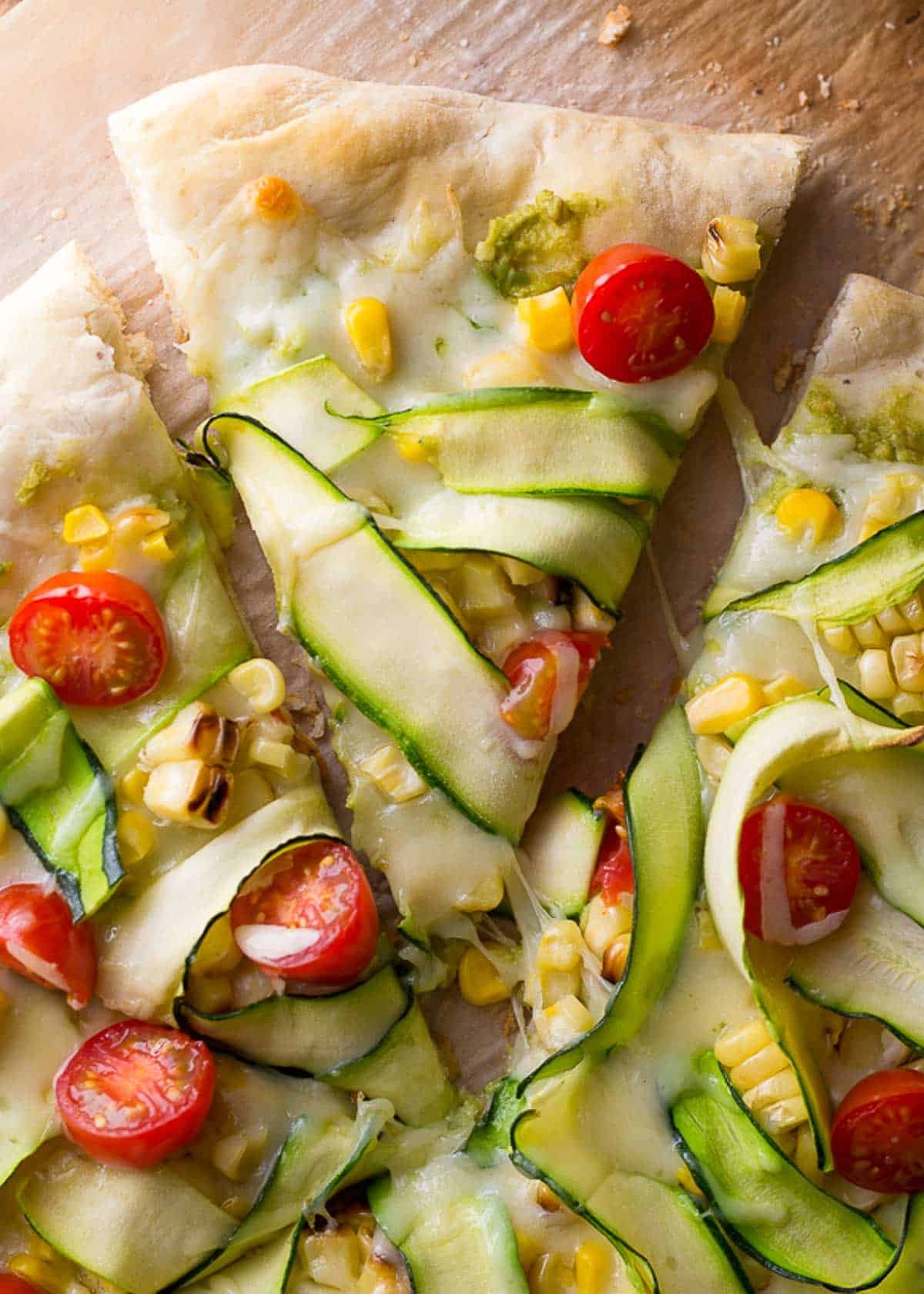 a slice of summer veggie pizza being pulled from the full flatbread pizza on a wooden table