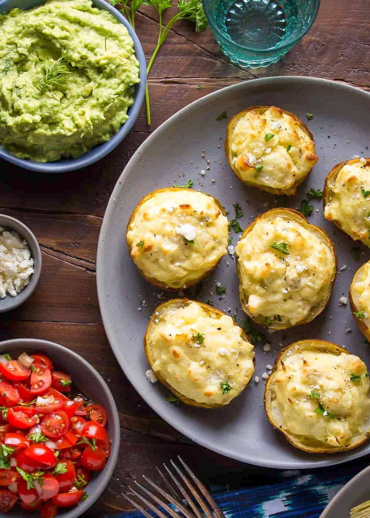 a plate of twice baked potatoes beside a bowl of avocado mash and a bowl of tomato bruschetta