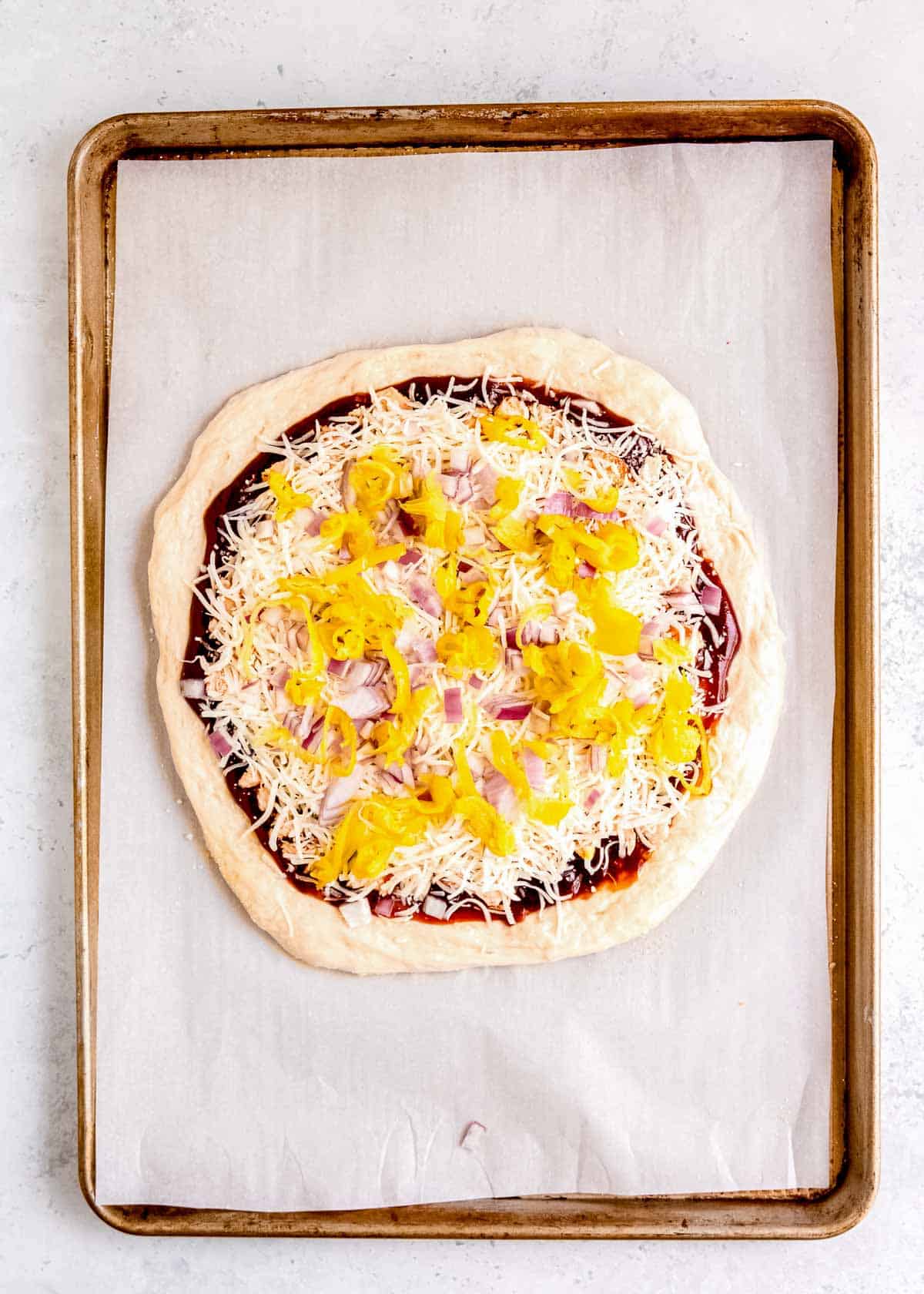 pizza dough topped with bbq sauce, pieces of chicken, shredded mozzarella, and tangy pepperoncini