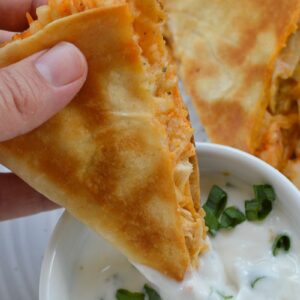 a slice of buffalo chicken quesadilla being dipped in sauce in a white bowl