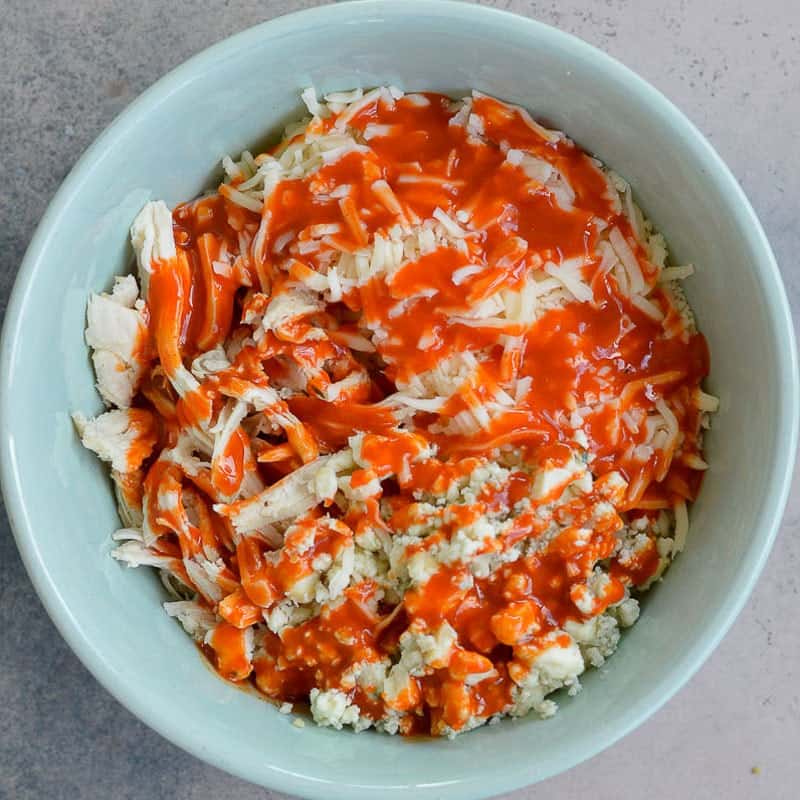 shredded chicken being mixed with buffalo sauce in a white bowl