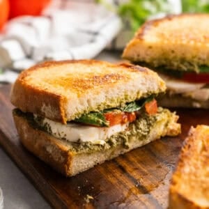 three pesto sandwiches on a cutting board with a white kitchen towel and additional ingredients in background