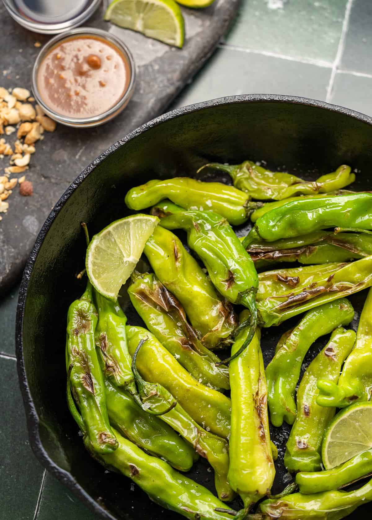 skillet full of blistered shishito peppers beside peanut sauce, peanuts, and lime wedges